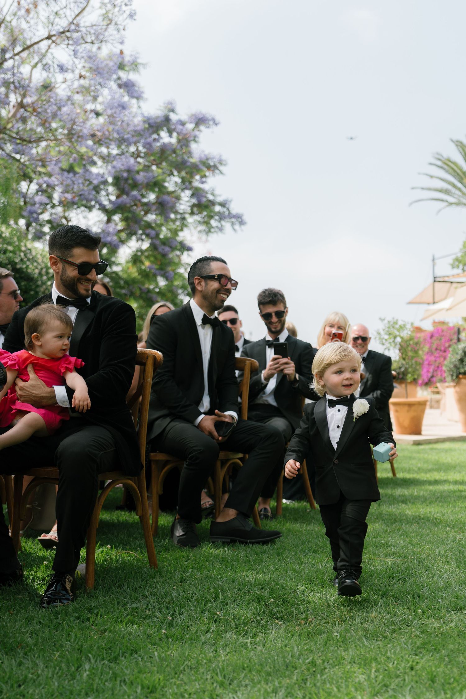 Wedding guests in formal attire seated on wooden chairs on a manicured lawn.