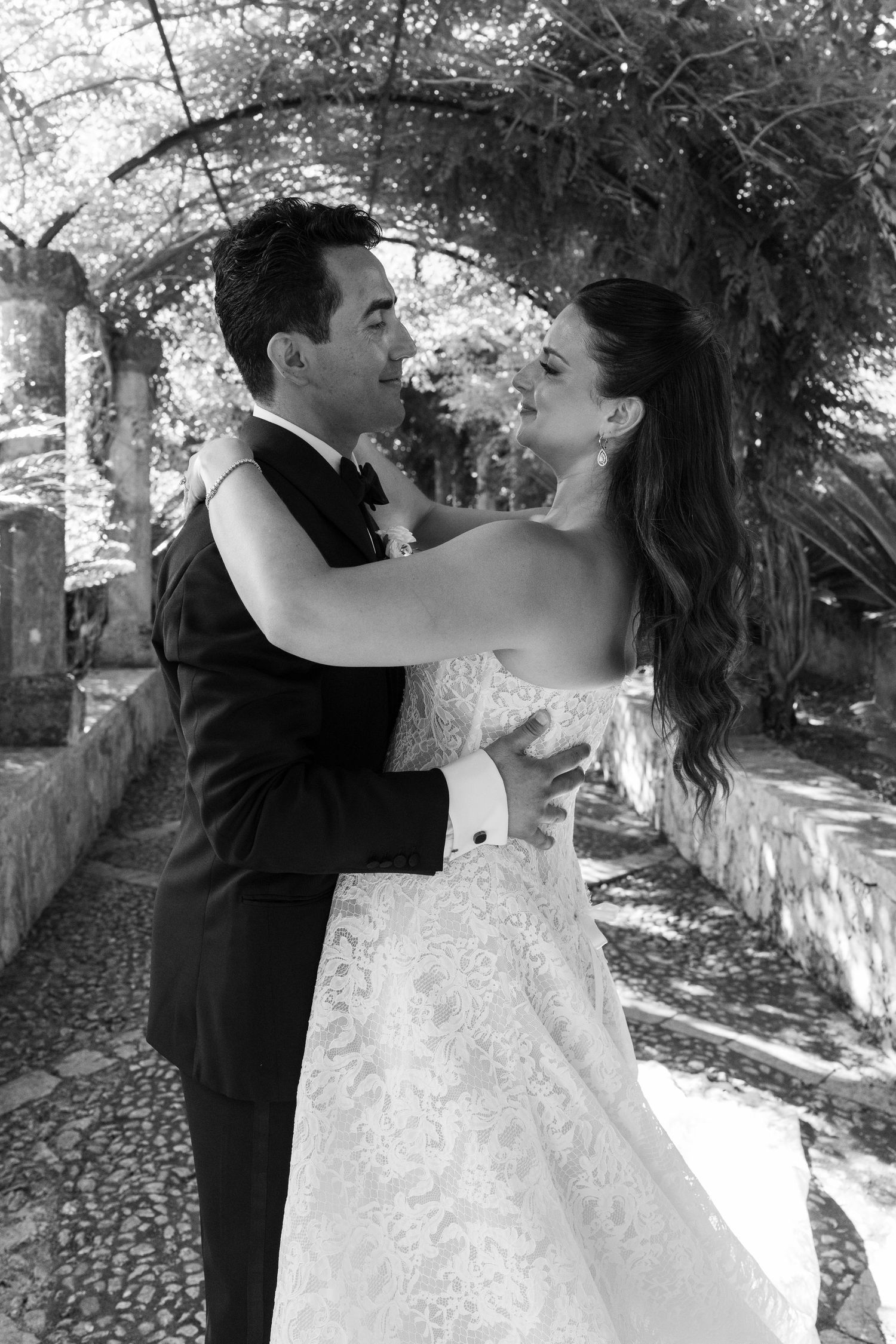 Black and white photo of a bride and groom embracing under a vine-covered archway in a garden setting.