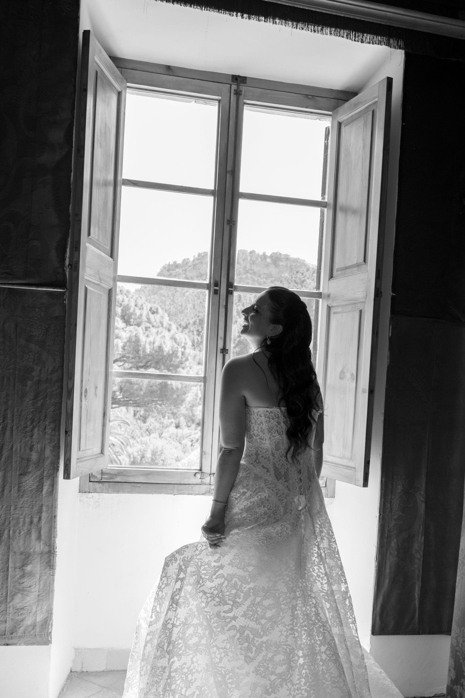 Black and white photo of a bride in lace gown gazing out an open window with scenic trees visible outside.