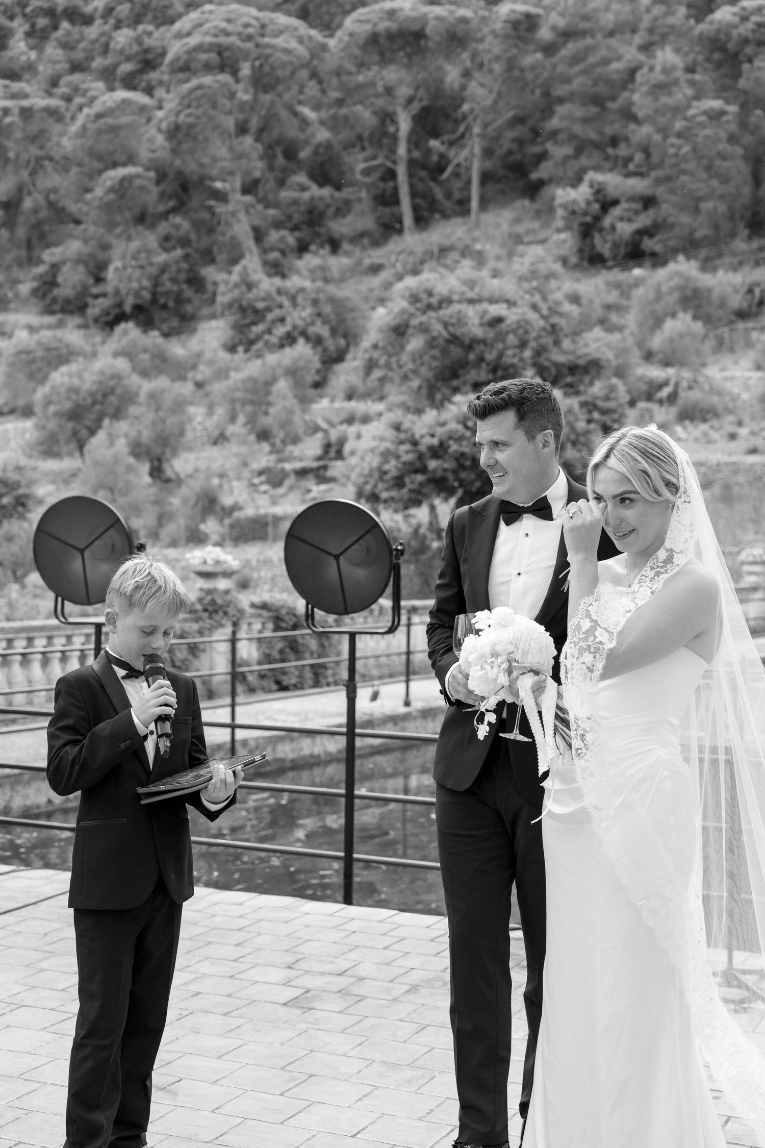Black and white wedding photo of bride and groom with young ring bearer reading vows outdoors.