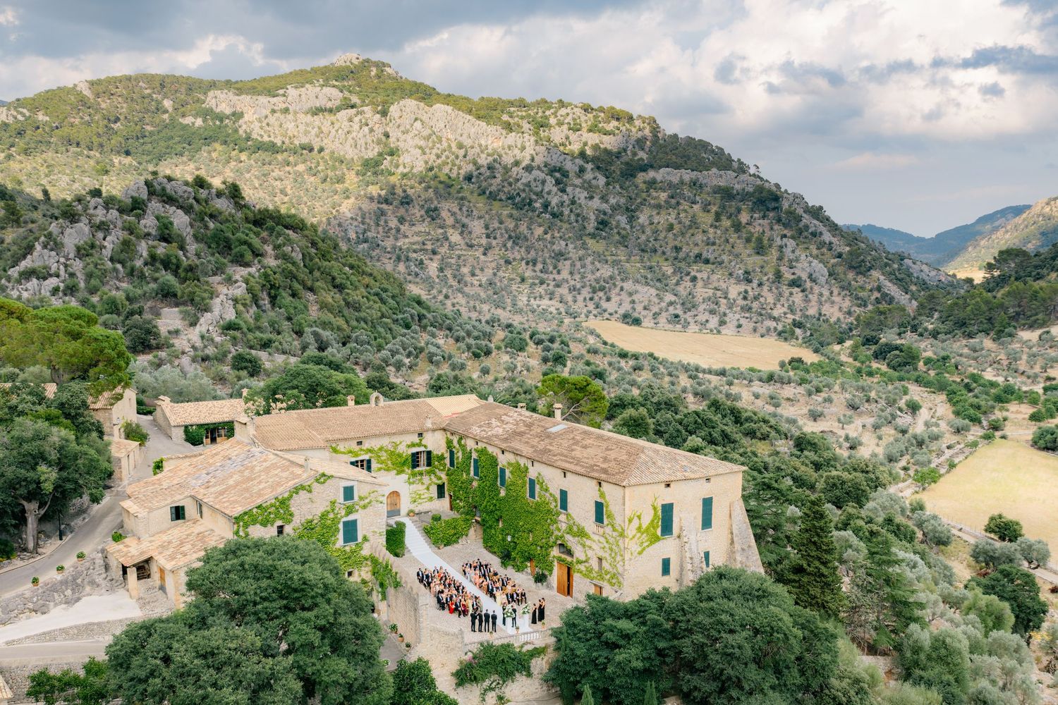 Aerial view of a rustic stone villa nestled in lush green mountains, with guests gathered in a courtyard.
