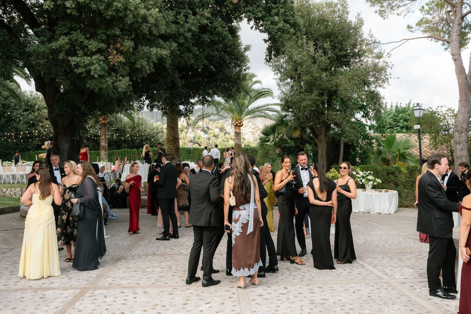 Elegant outdoor wedding reception with guests mingling in formal attire surrounded by lush trees.