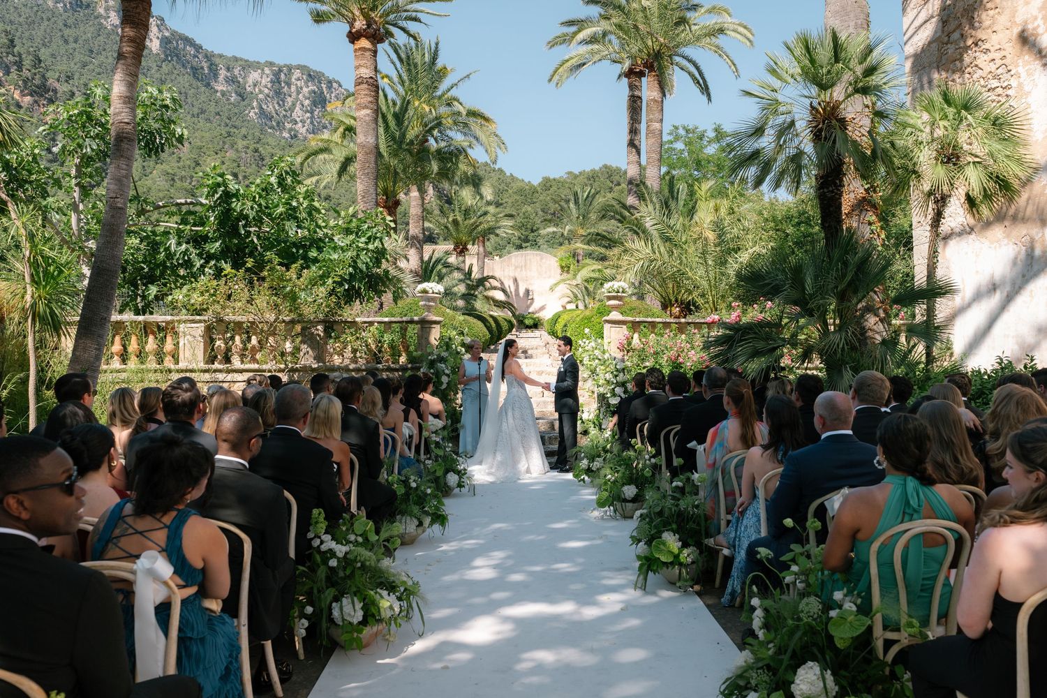 Elegant outdoor wedding ceremony with white aisle, lush greenery, and palm trees at a Mediterranean villa.