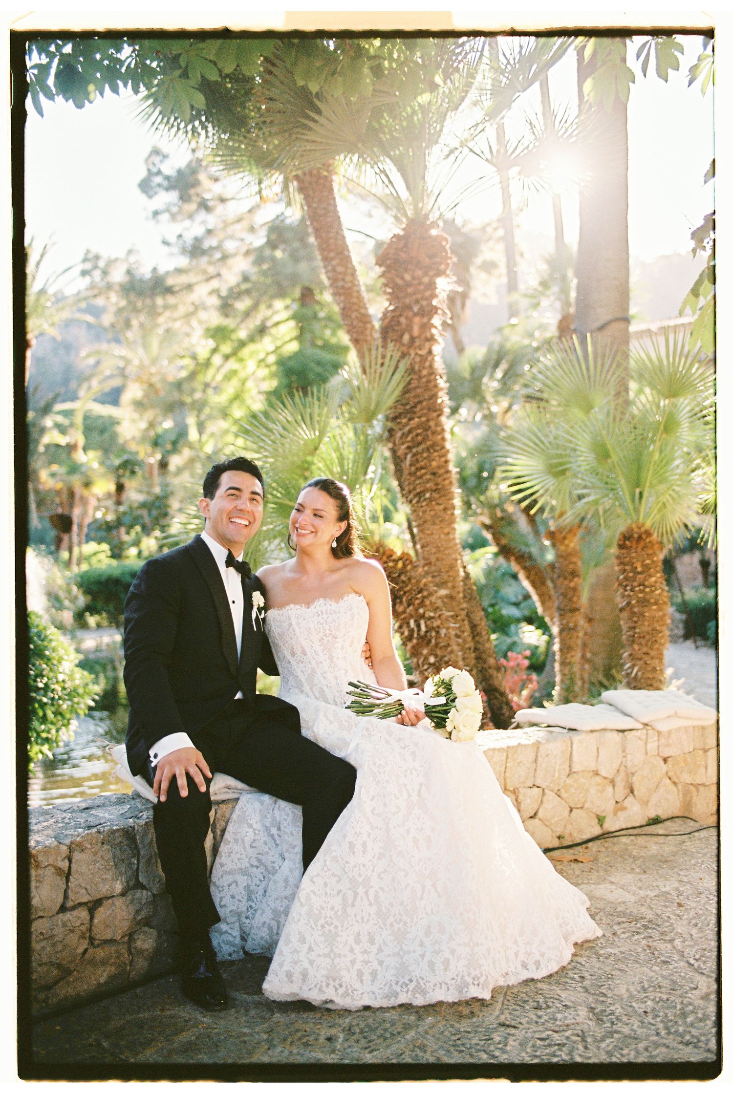 Bride and groom sitting on stone wall surrounded by palm trees in lush garden setting on their wedding day.