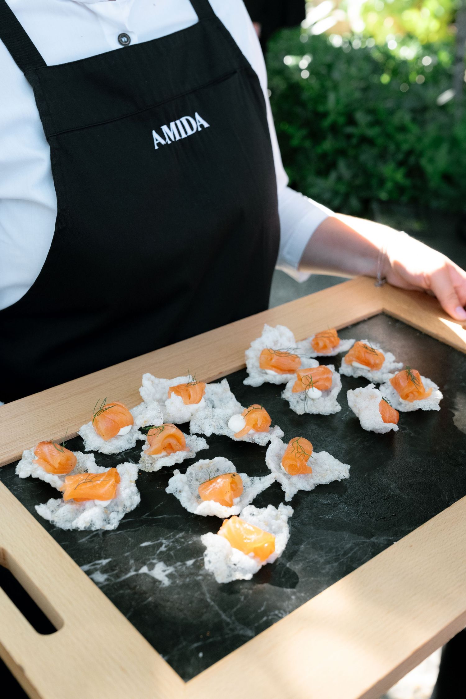 AMIDA chef holding a wooden tray with elegant smoked salmon canapes on a black marble slate.