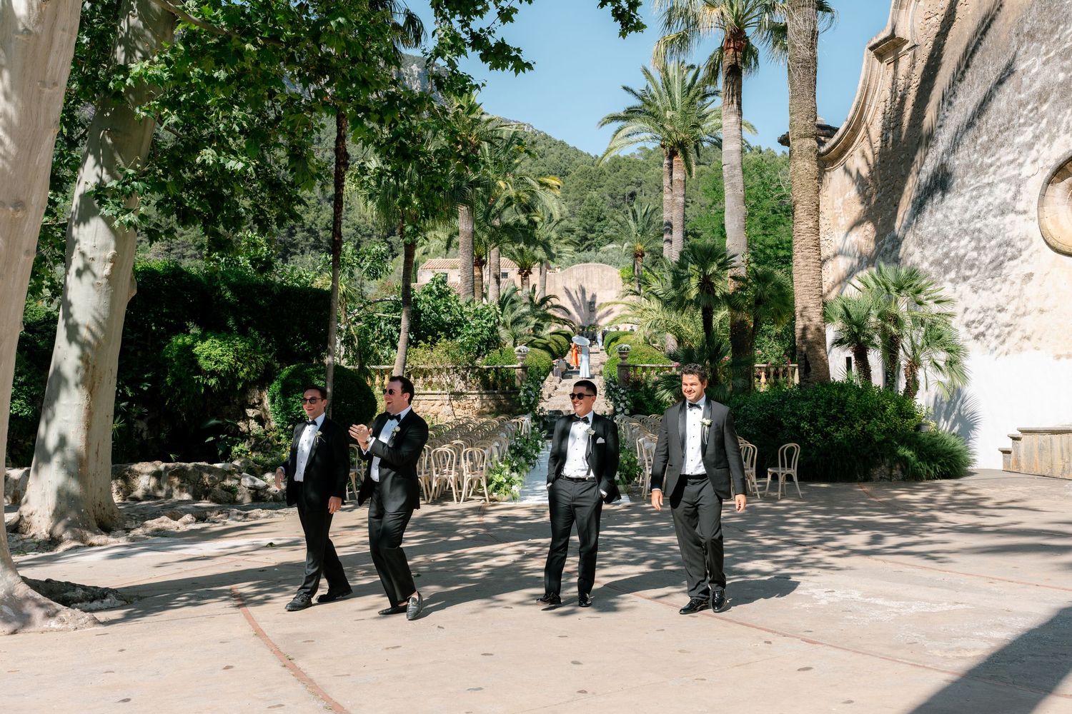 Groomsmen in black tuxedos celebrate outdoors near a stone building surrounded by palm trees and lush greenery.