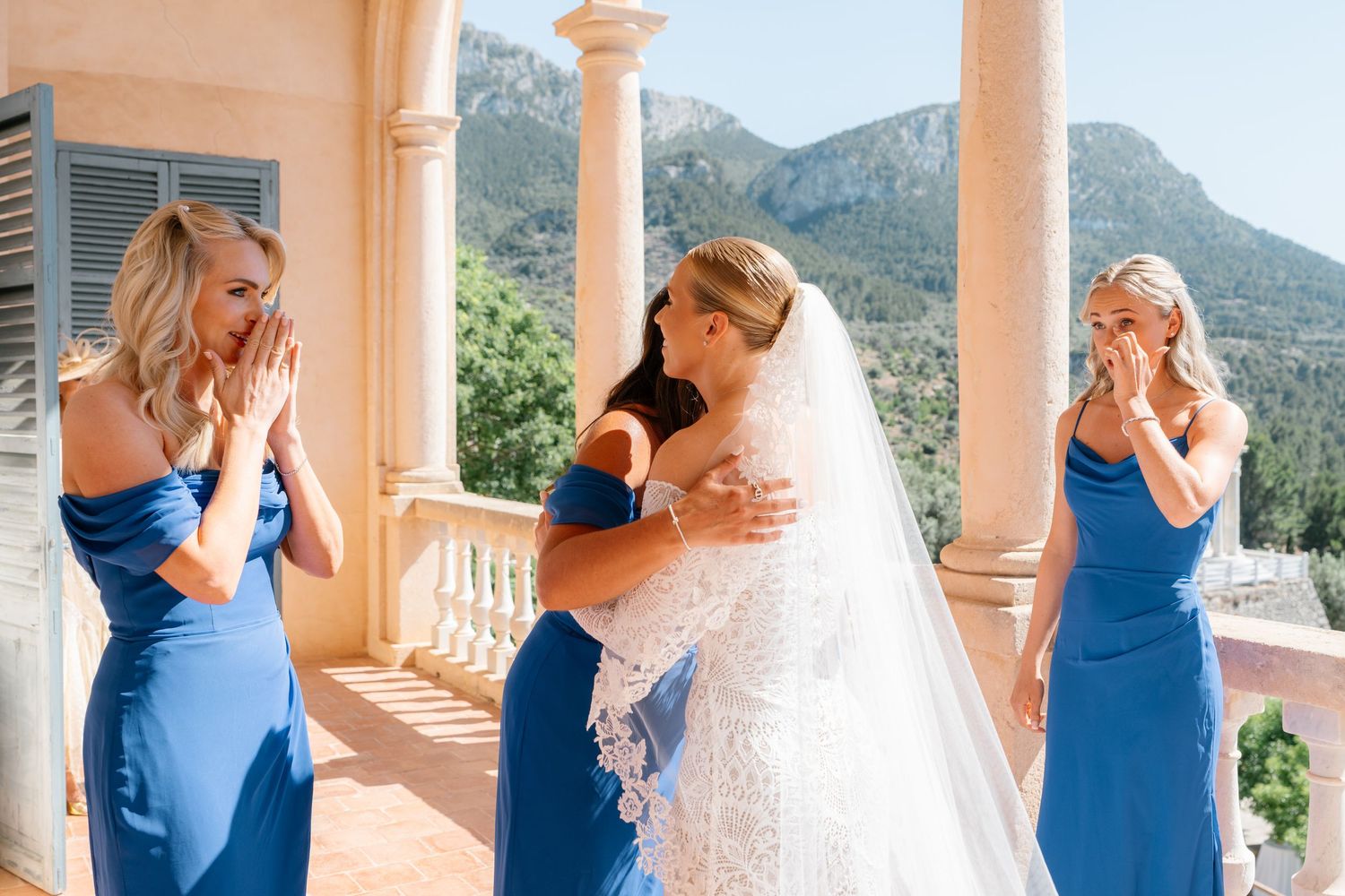 Wedding party in blue bridesmaid dresses gathered on a sunny terrace with mountain backdrop.