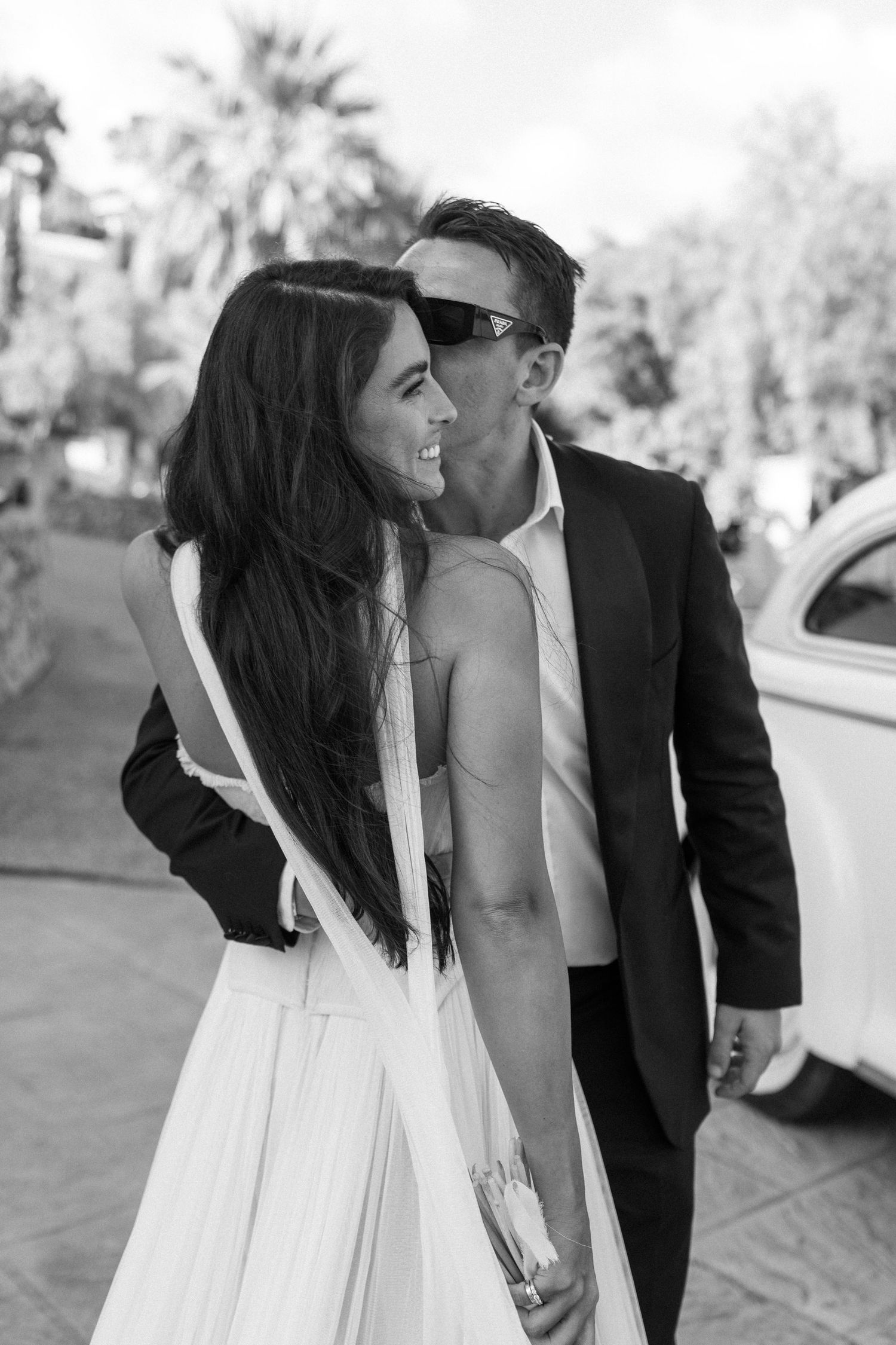 A bride in a white dress and groom in black suit share a candid laughing moment together outdoors.