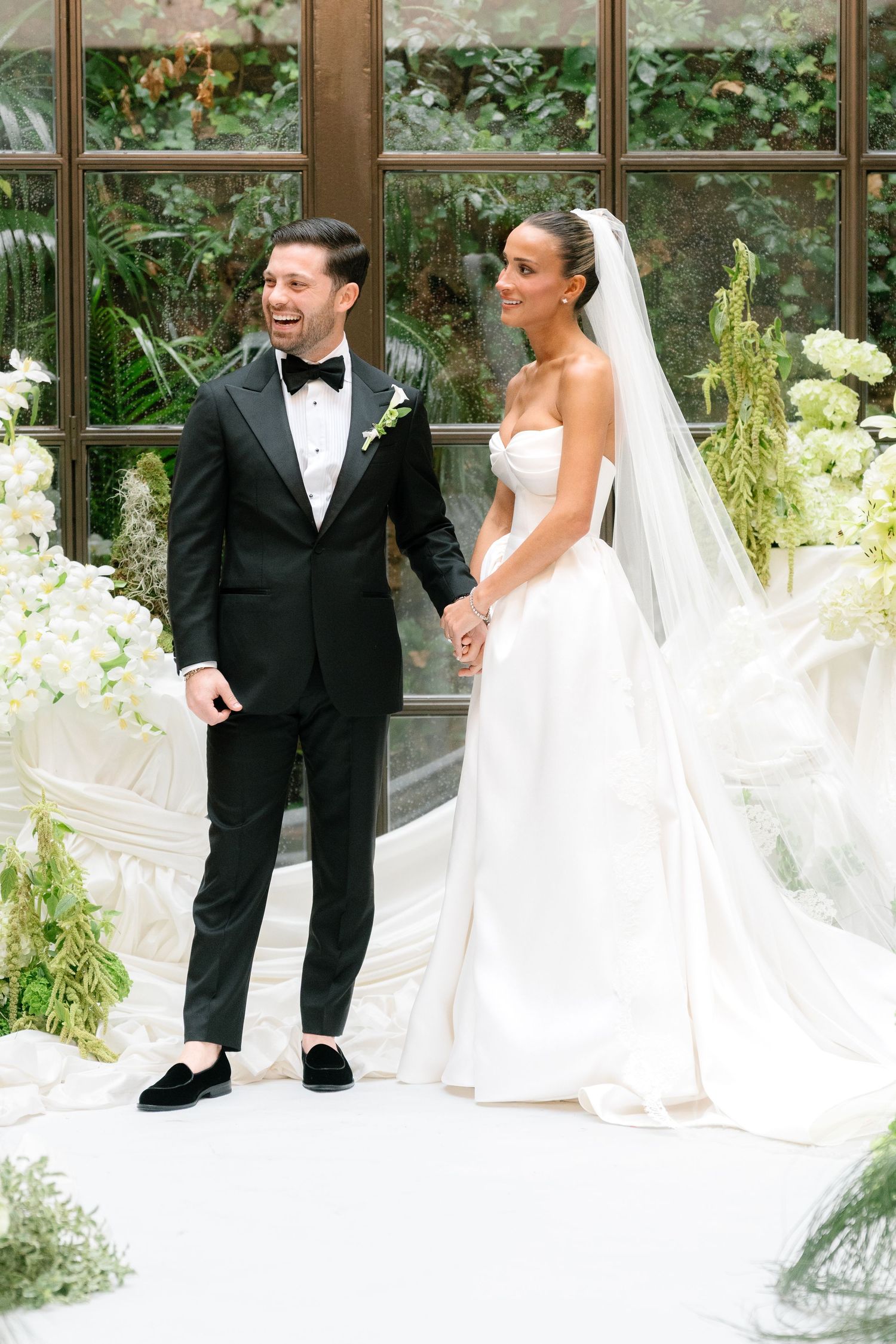 Elegant wedding couple in formal attire walking through greenhouse venue with white floral decorations.