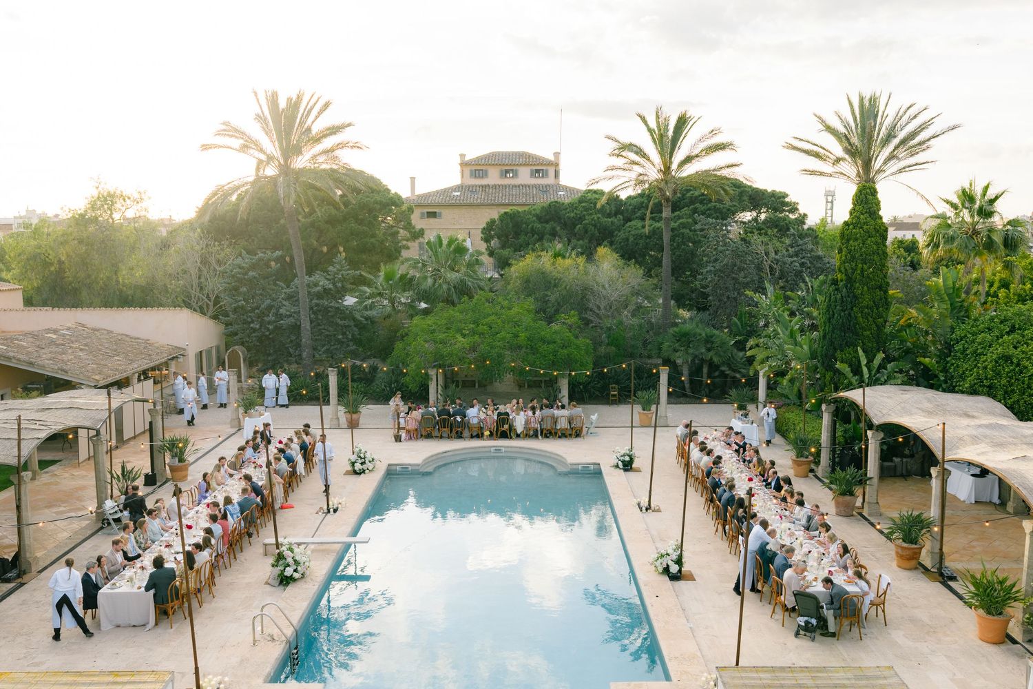 Aerial view of a resort venue with palm trees and swimming pools surrounded by stone pathways.