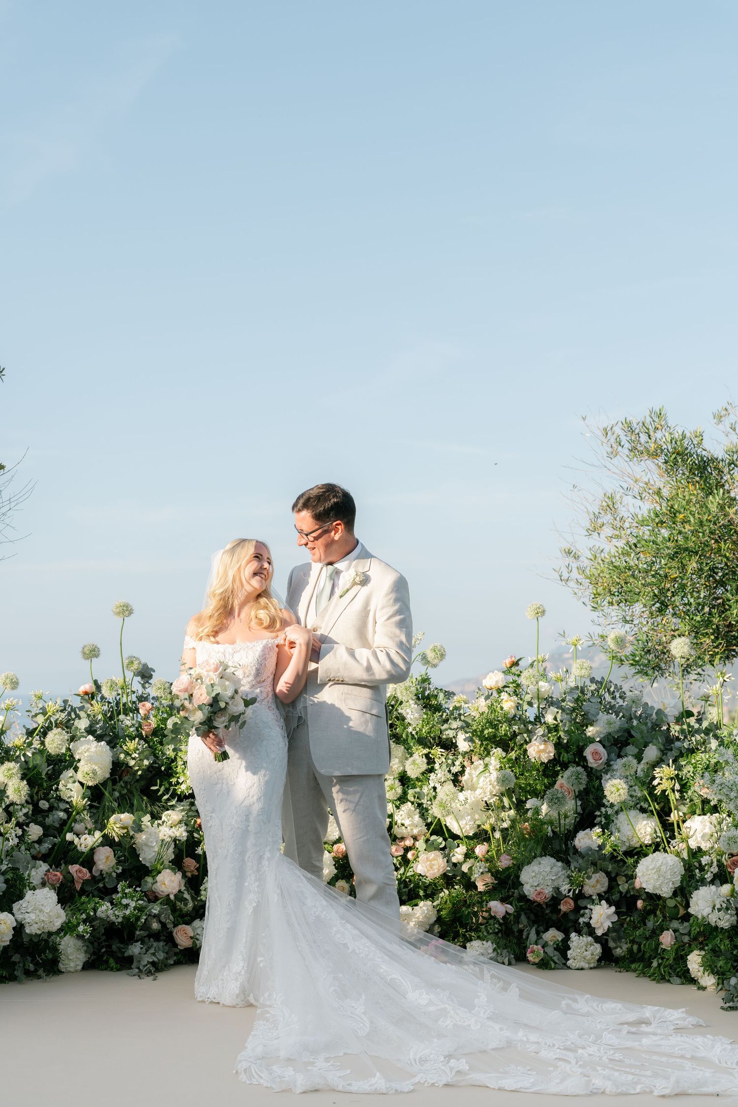 Wedding couple posing against white floral arch with mountain backdrop.