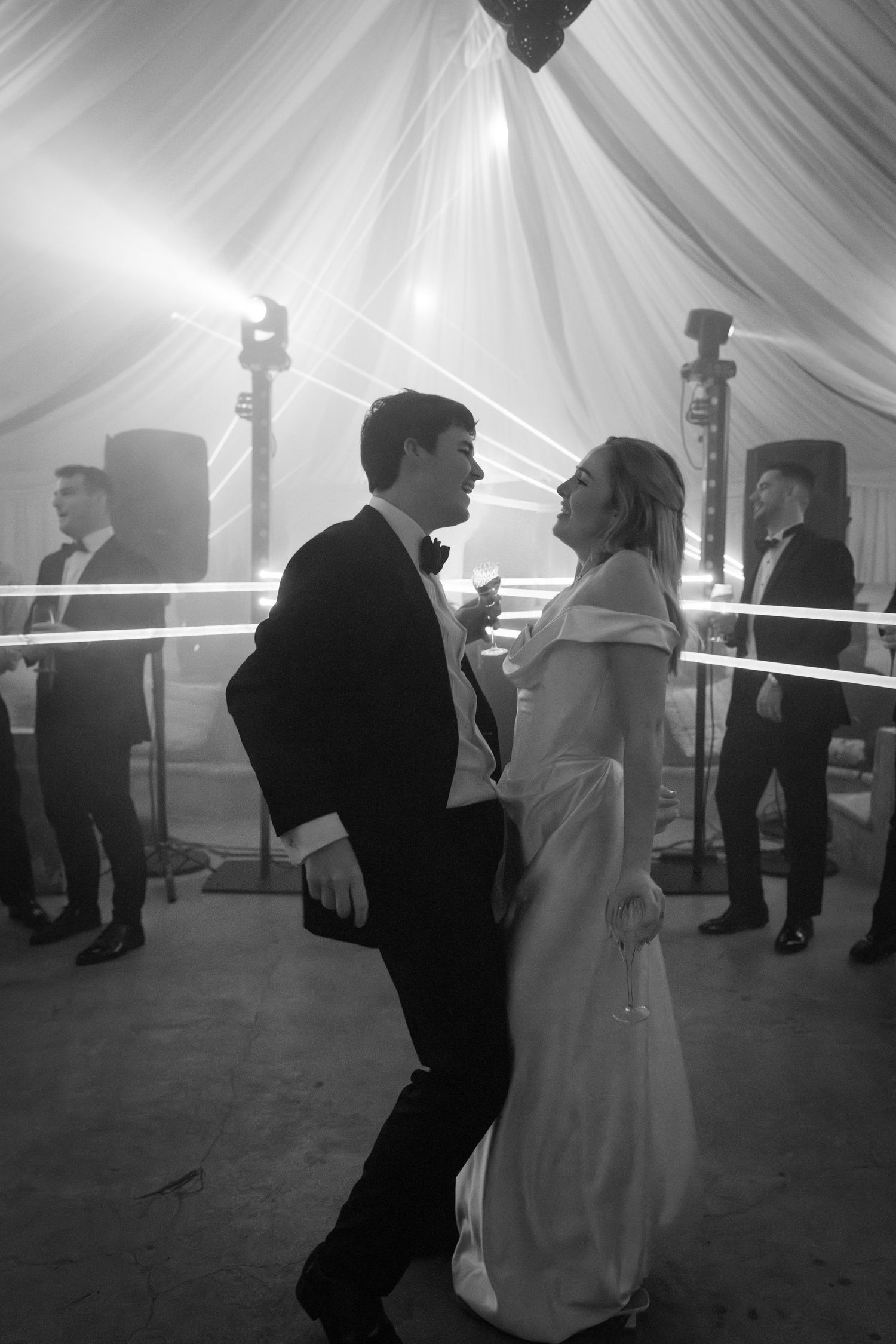 Black and white photo of couples dancing in a draped tent reception space with dramatic lighting.