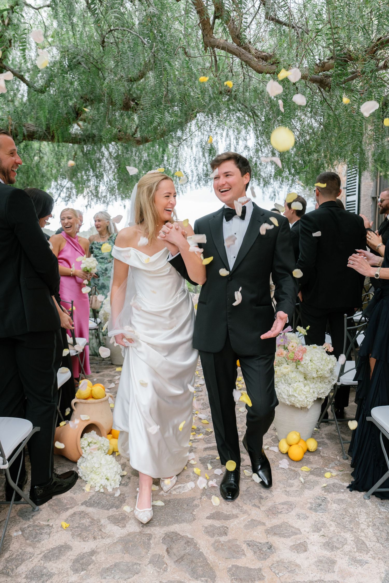 Wedding celebration on stone patio with guests in formal attire dancing under willow trees.