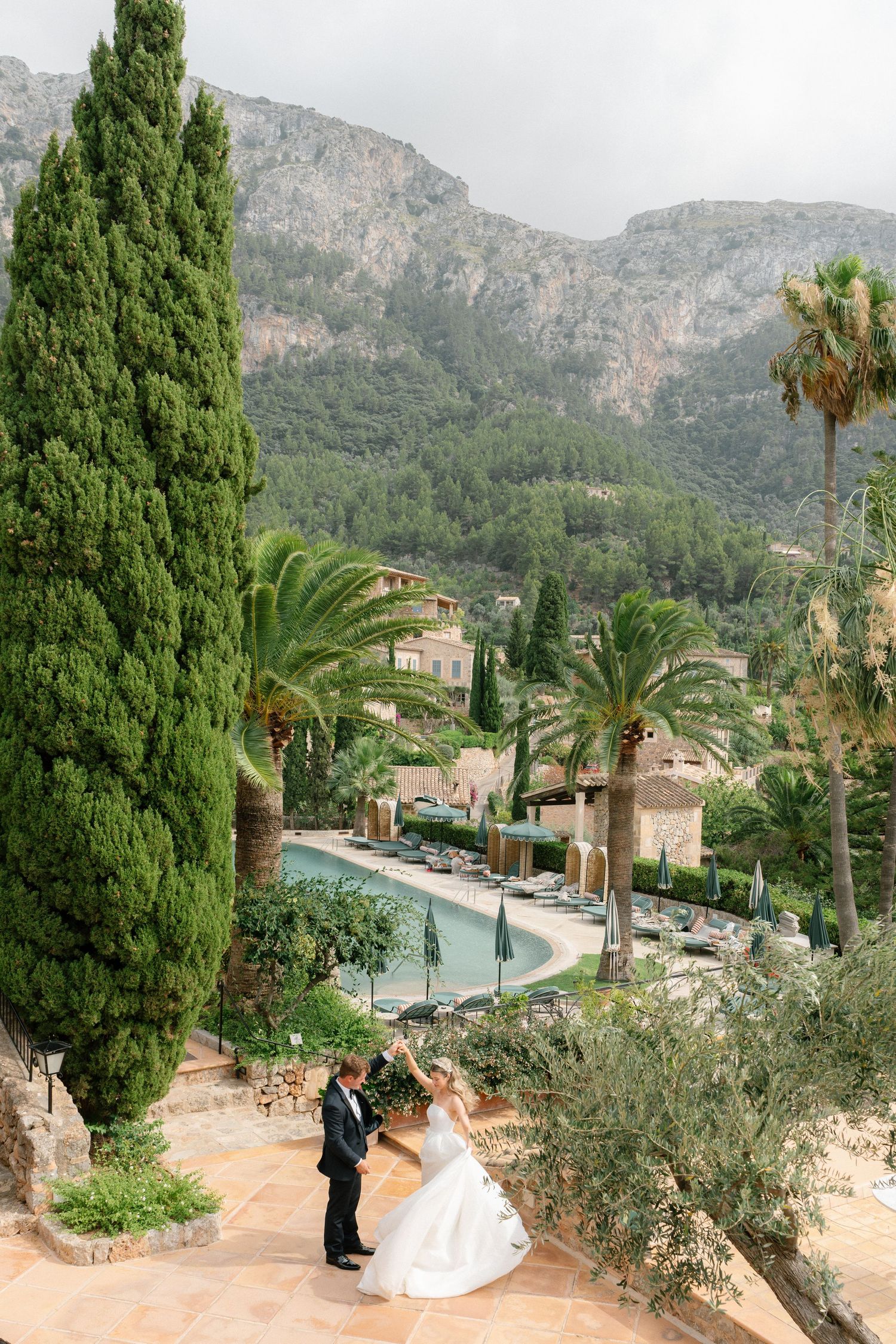 A wedding couple poses on stone steps overlooking a Mediterranean villa with mountains and cypress trees in the background.