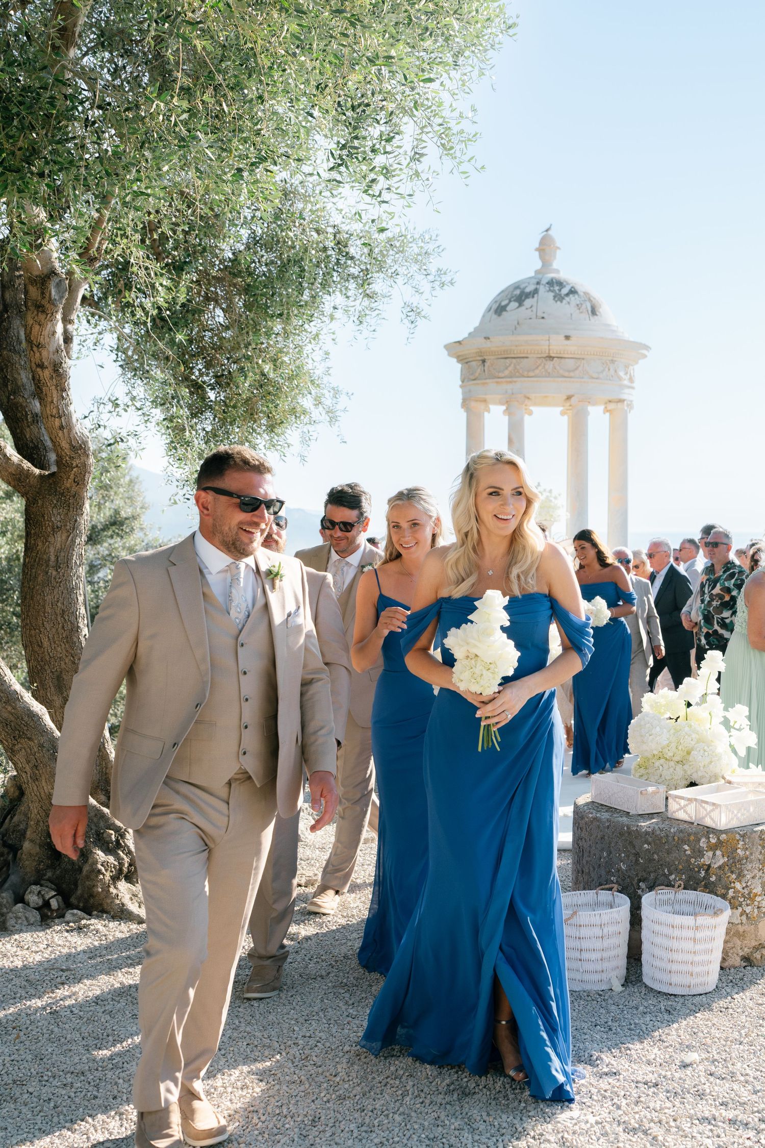 Wedding party in beige suits and blue dresses walking outdoors near a gazebo.