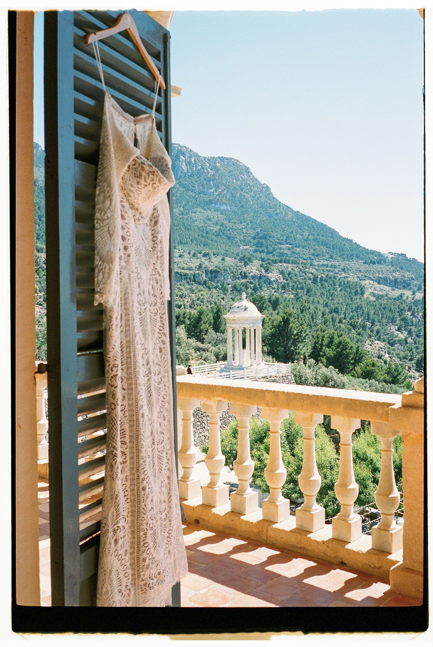 Elegant lace wedding dress hangs against a Mediterranean backdrop with mountain views and stone balustrade.