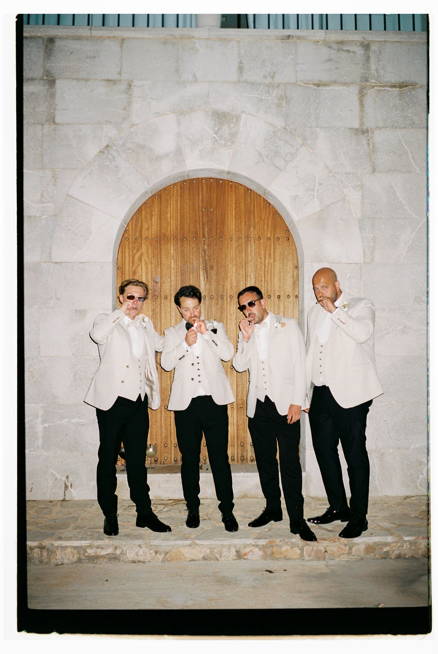 Four groomsmen in matching black and white attire pose playfully against a white wall with wooden door.
