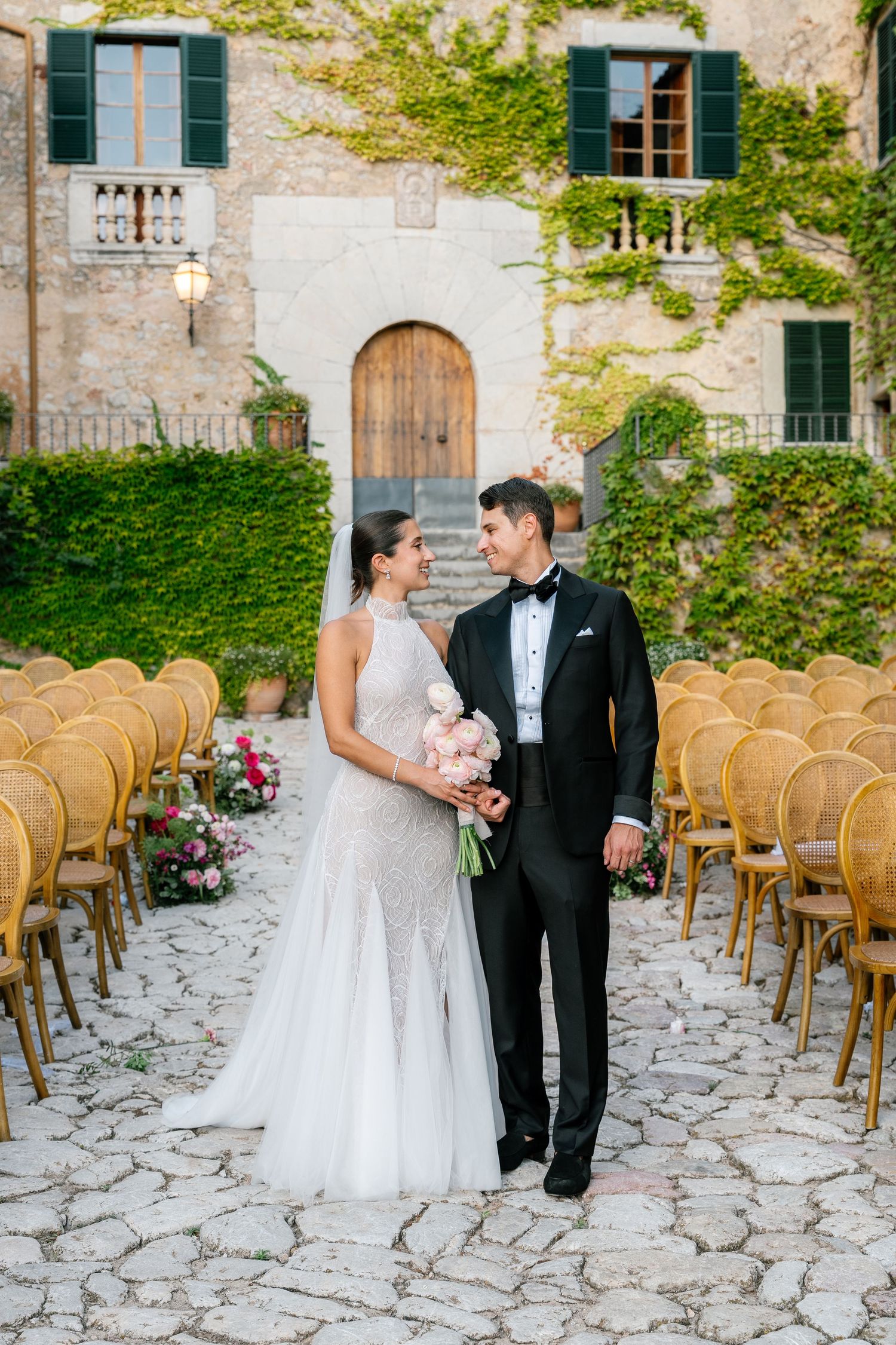 Newlyweds stand in an outdoor ceremony space with wooden chairs and stone architecture.