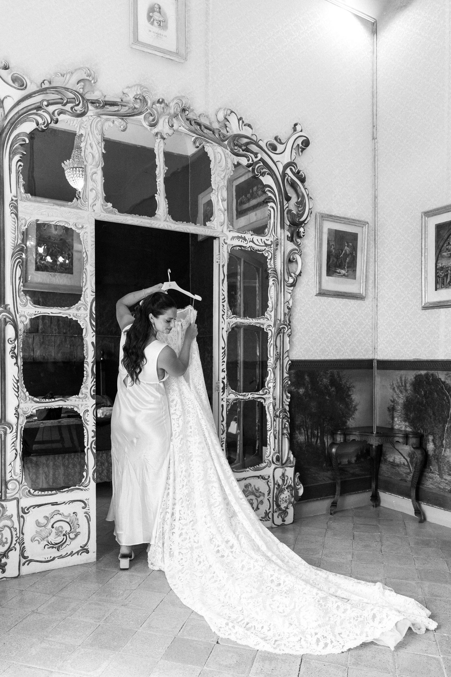 Black and white photo of a bride in a long white gown with lace train standing before an ornate baroque mirror.