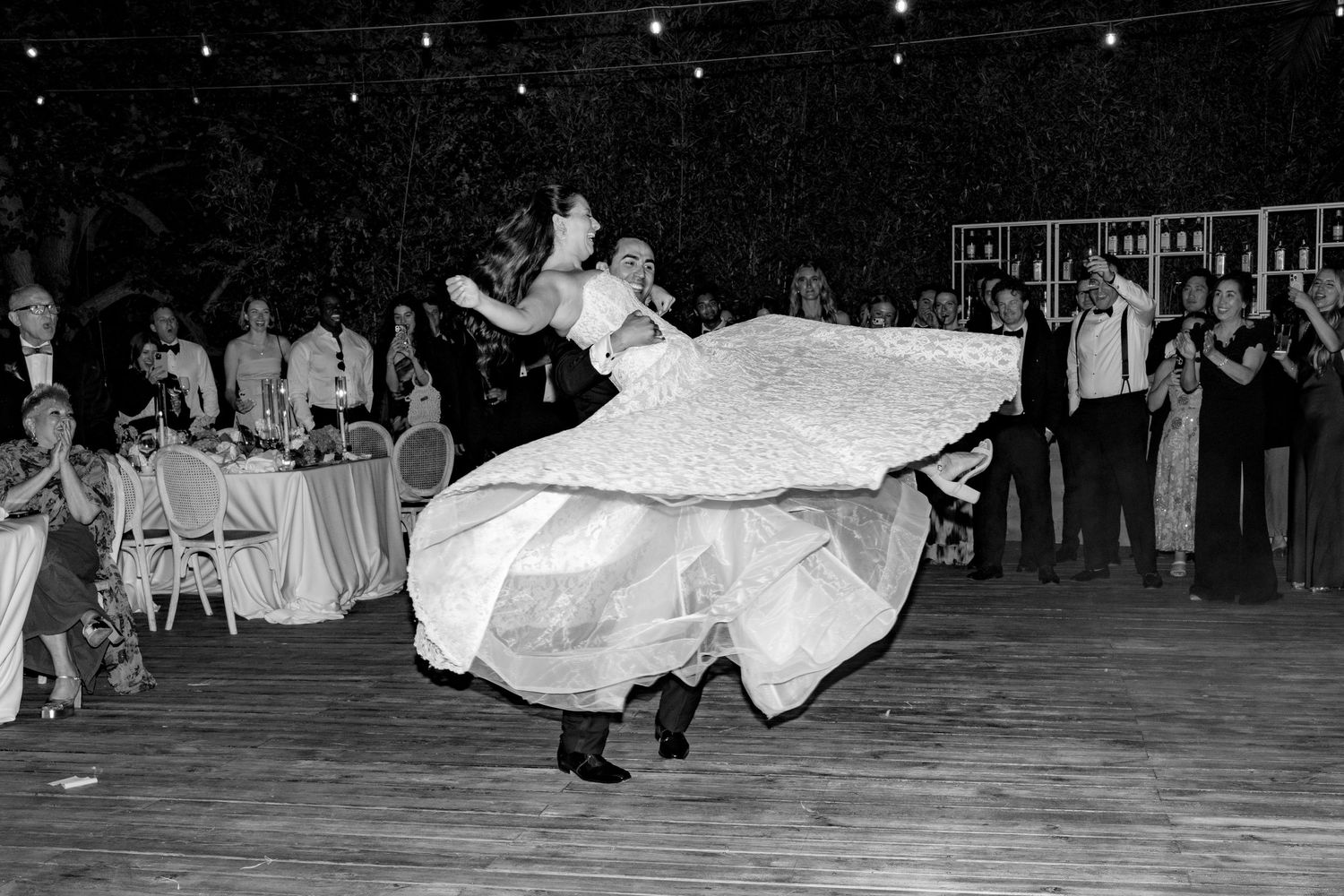 Black and white photo of bride and groom dancing energetically, wedding dress swirling, guests watching in background.