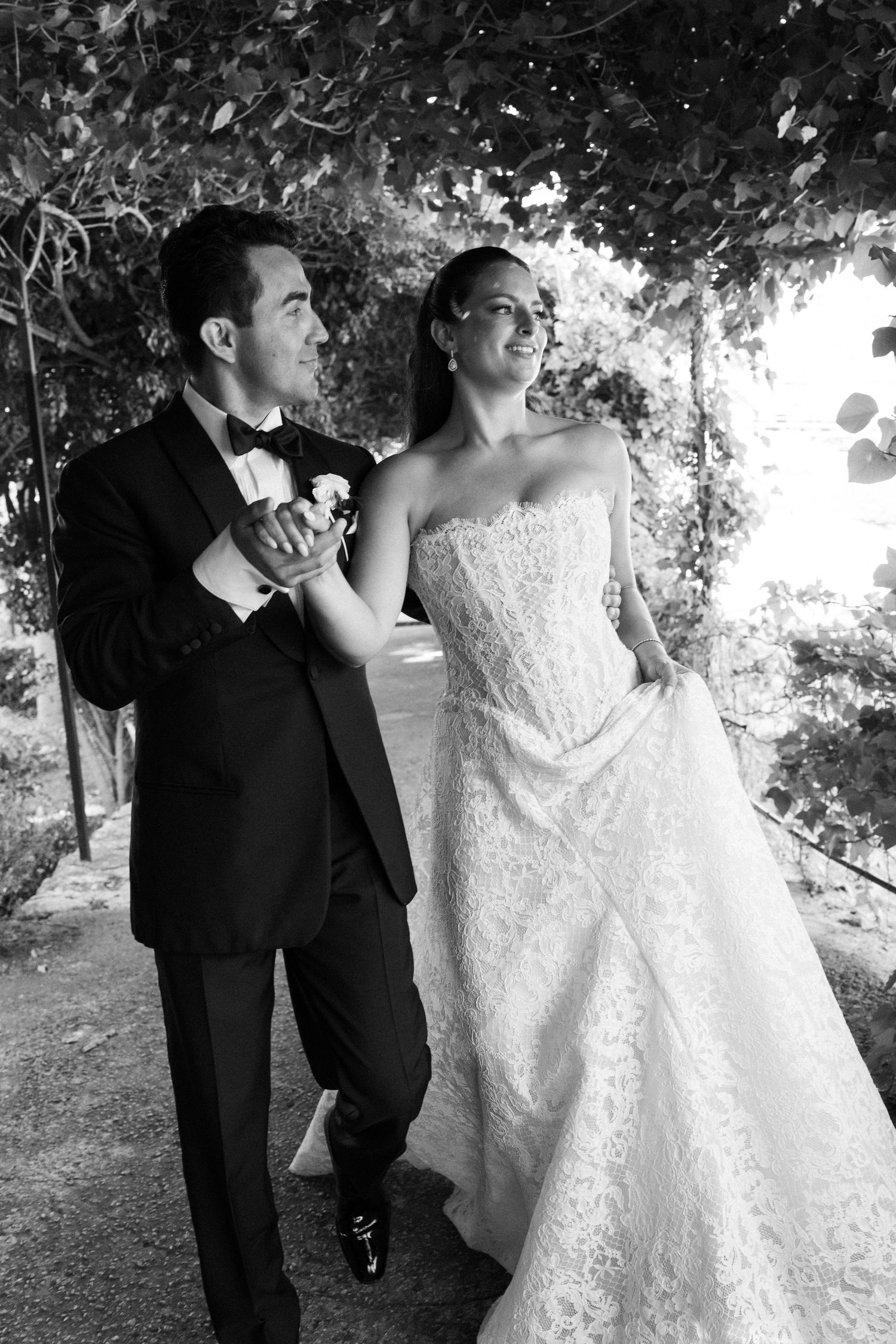 Black and white photo of a bride and groom holding hands under a leafy canopy on their wedding day.