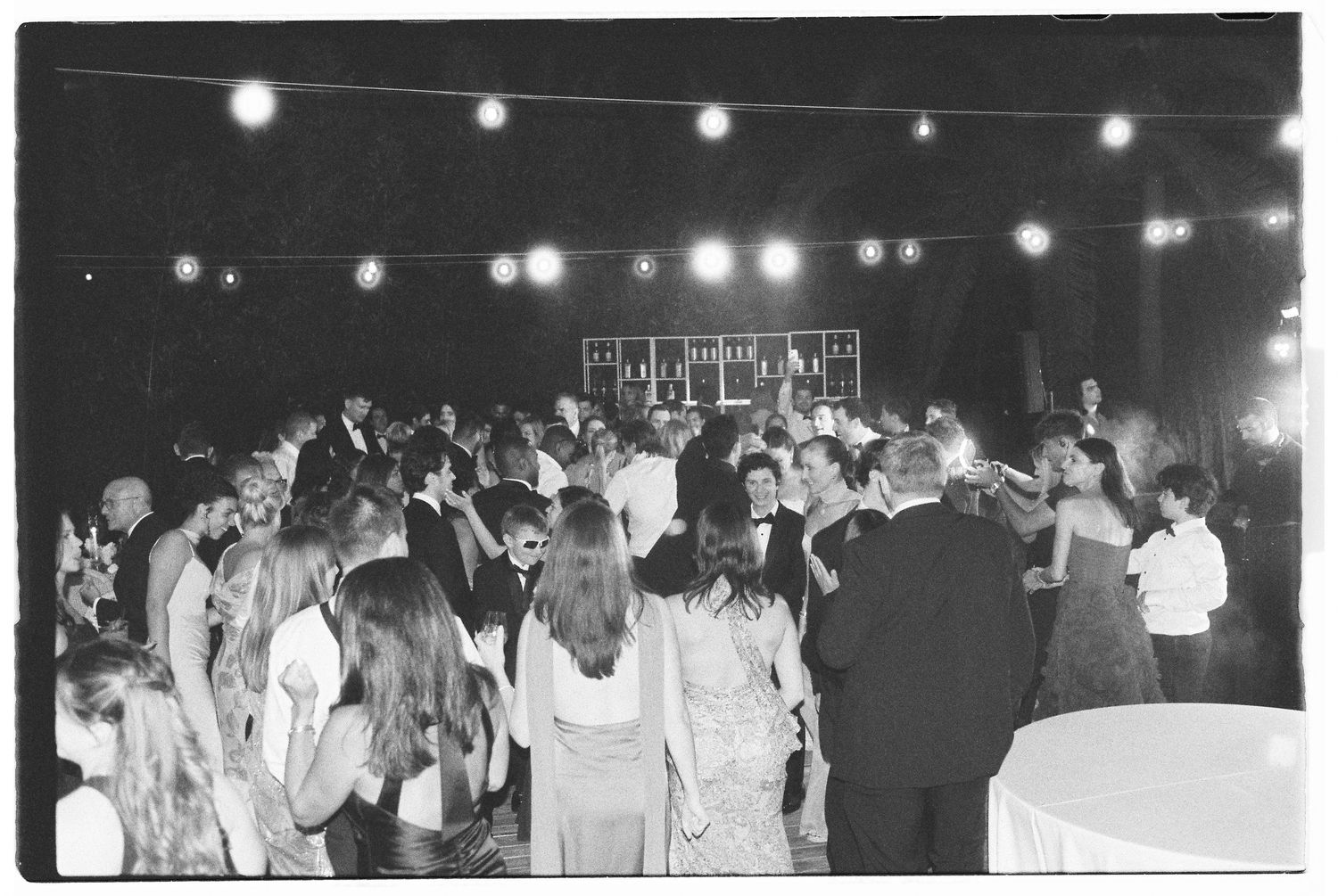 Black and white photo of a crowded wedding reception dance floor with string lights overhead.