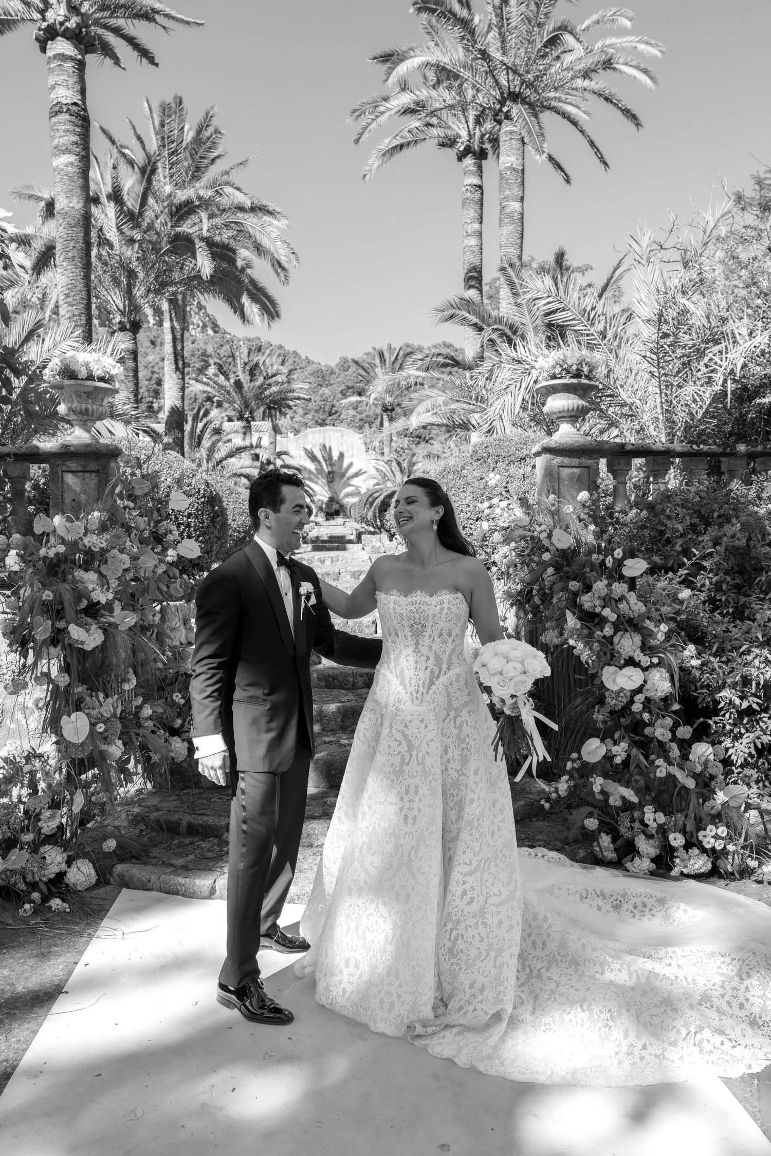 Black and white photo of a bride and groom walking among palm trees and floral arrangements at an outdoor wedding venue.