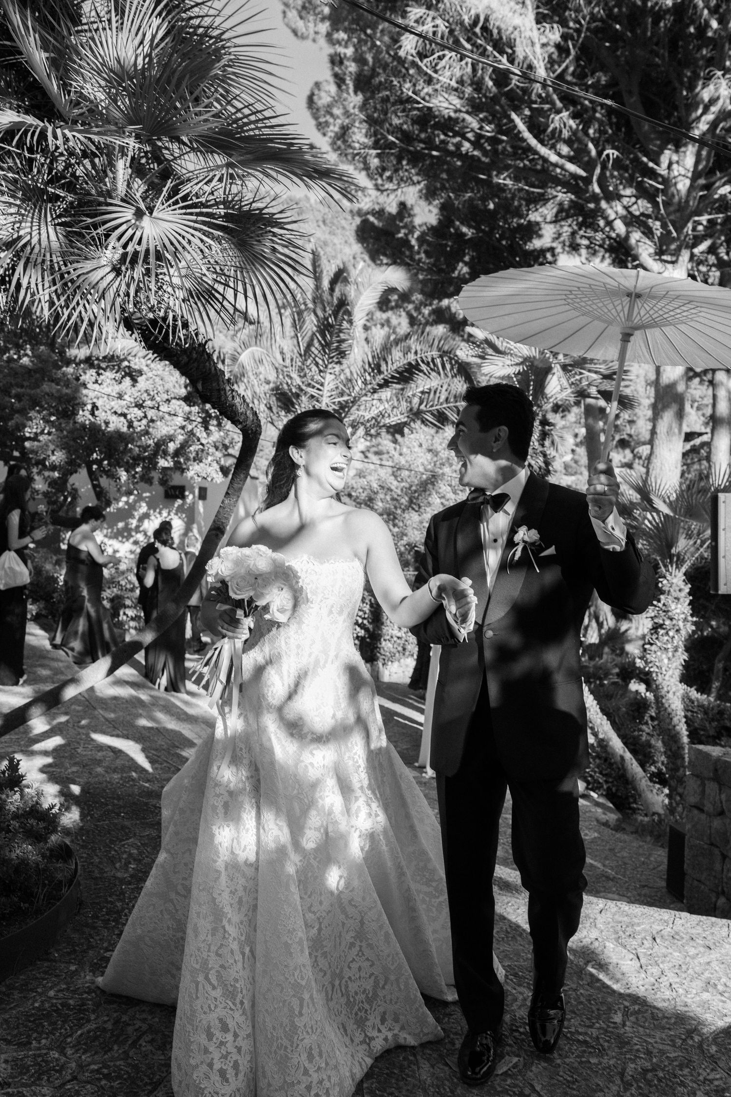 Black and white photo of a bride and groom walking together under a parasol in a lush garden setting.