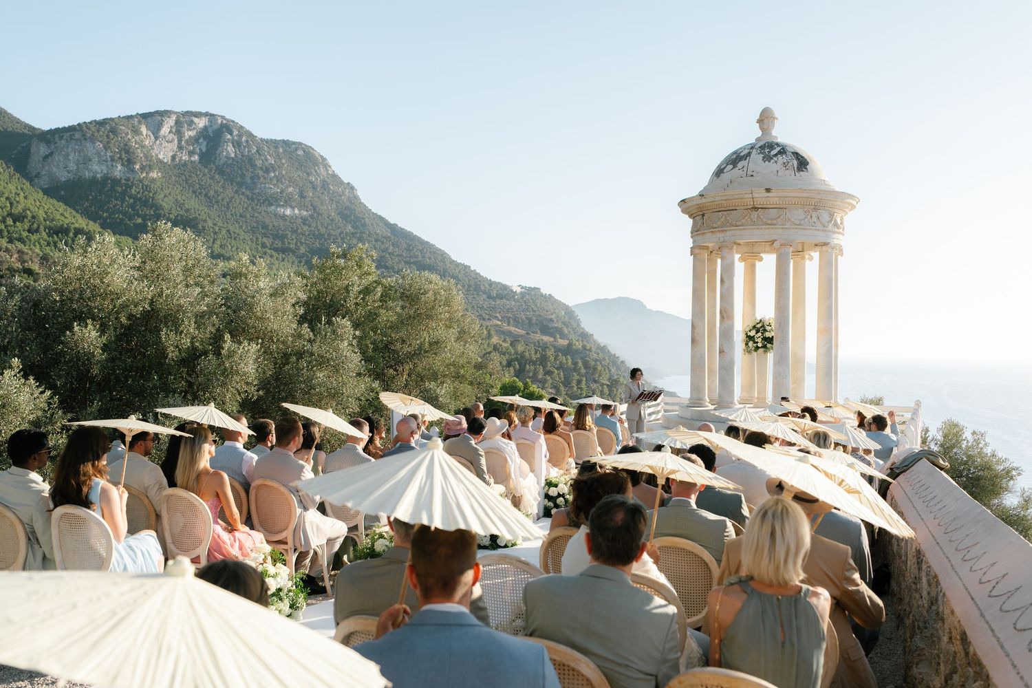 Outdoor wedding ceremony on a cliffside terrace in Mallorca with white umbrellas, guests seated, and a classical temple backdrop.