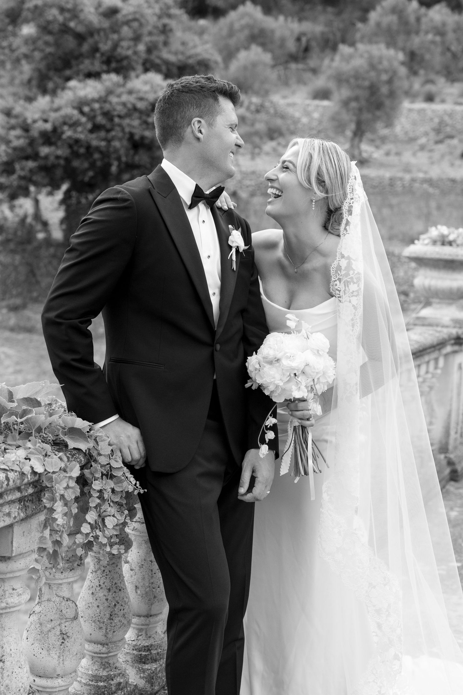 Black and white photo of smiling bride and groom in elegant wedding attire, sharing a joyful moment outdoors.