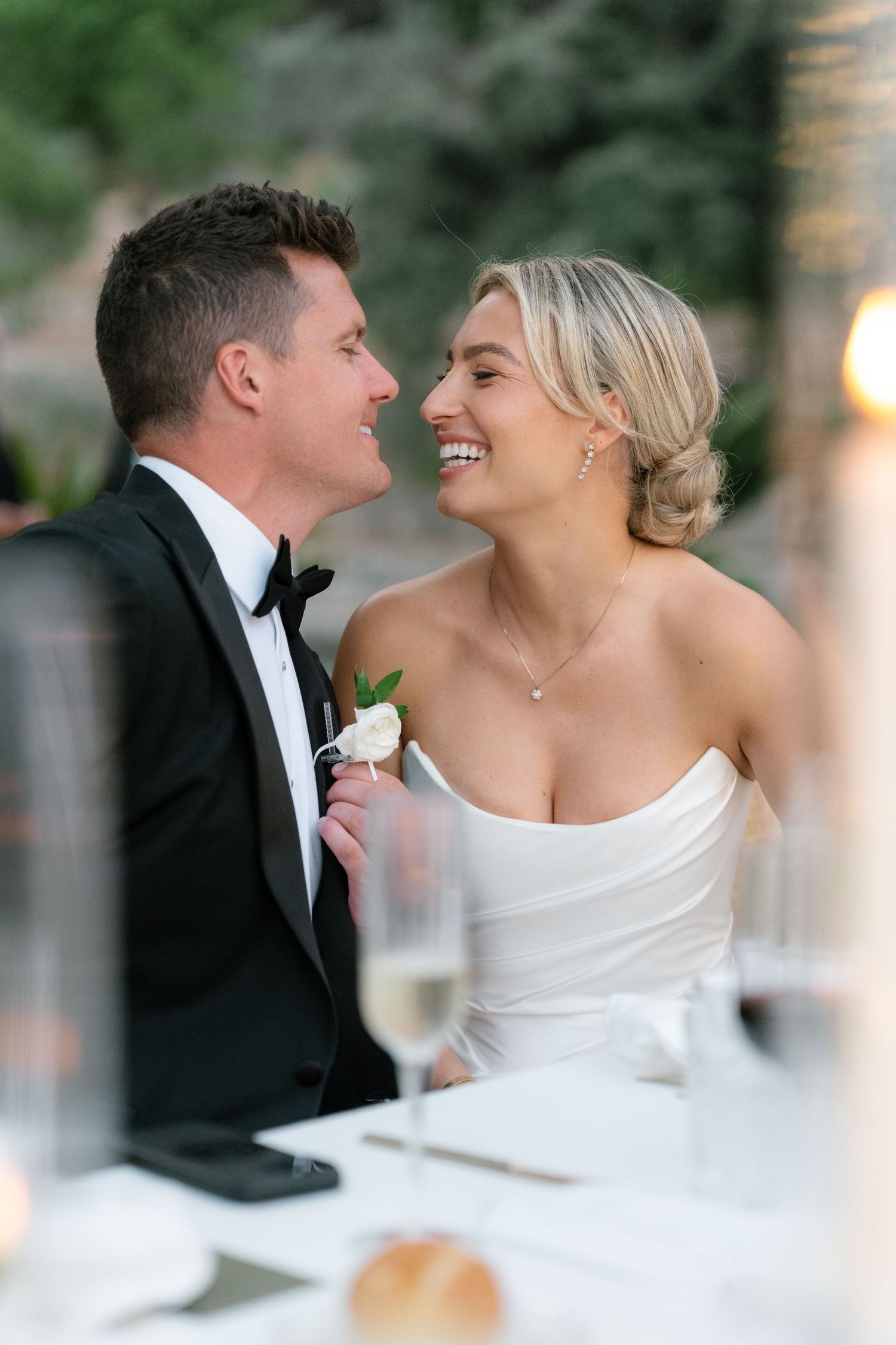 A smiling bride and groom in formal wedding attire share a romantic moment at their elegant reception dinner table.