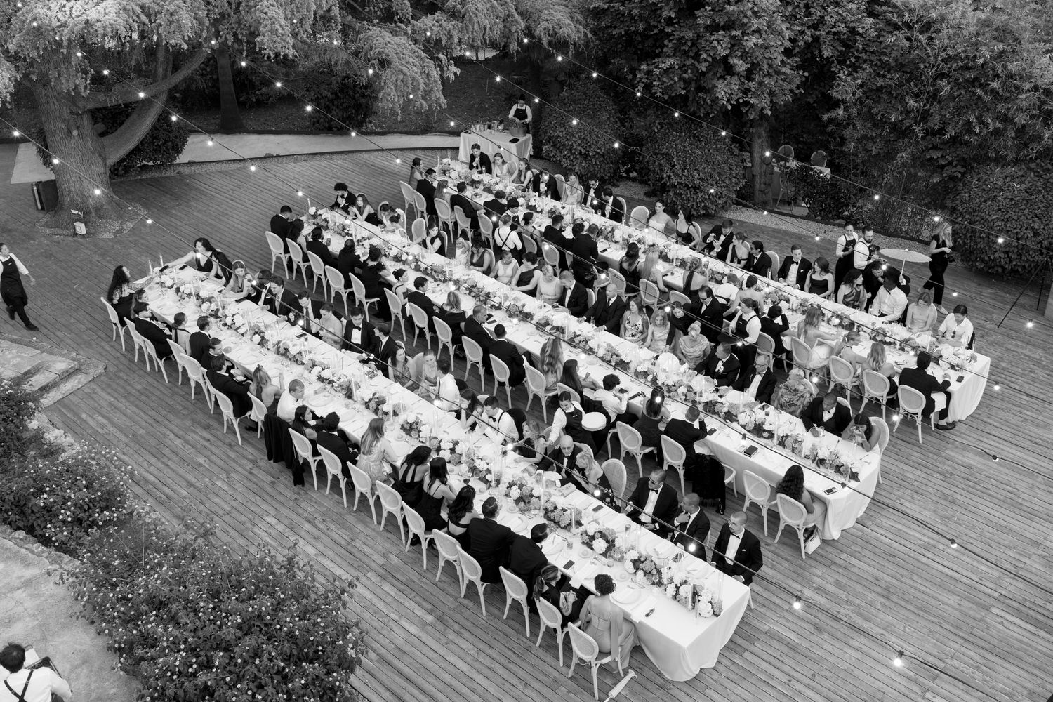 Aerial black and white photo of a large outdoor wedding reception with long banquet tables on a wooden deck.