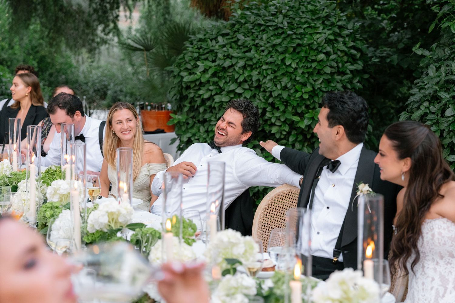 Guests laughing and celebrating at an elegant outdoor wedding reception with white floral arrangements and candlelit long table.