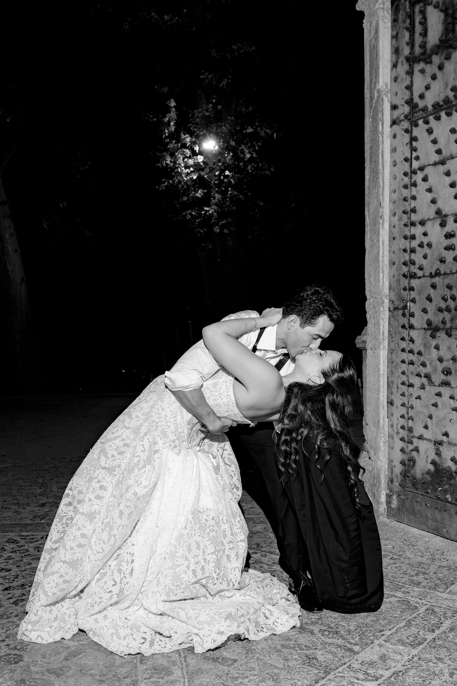 Black and white photo of a couple sharing a romantic dip kiss in wedding attire outside at night.