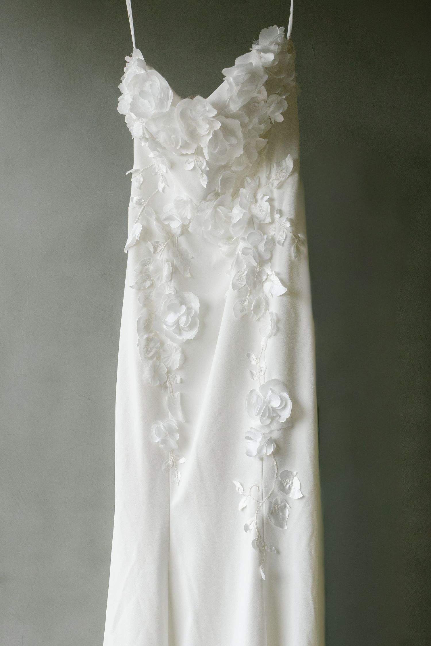 Close-up of white wedding dress with delicate floral appliques hanging against dark background.