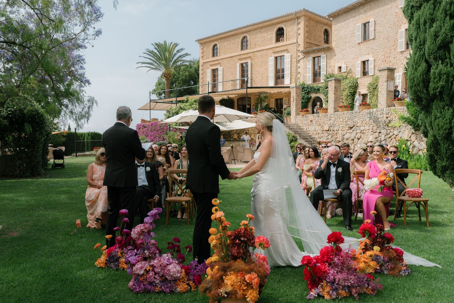 Outdoor wedding ceremony at elegant stone villa with colorful floral arrangements.