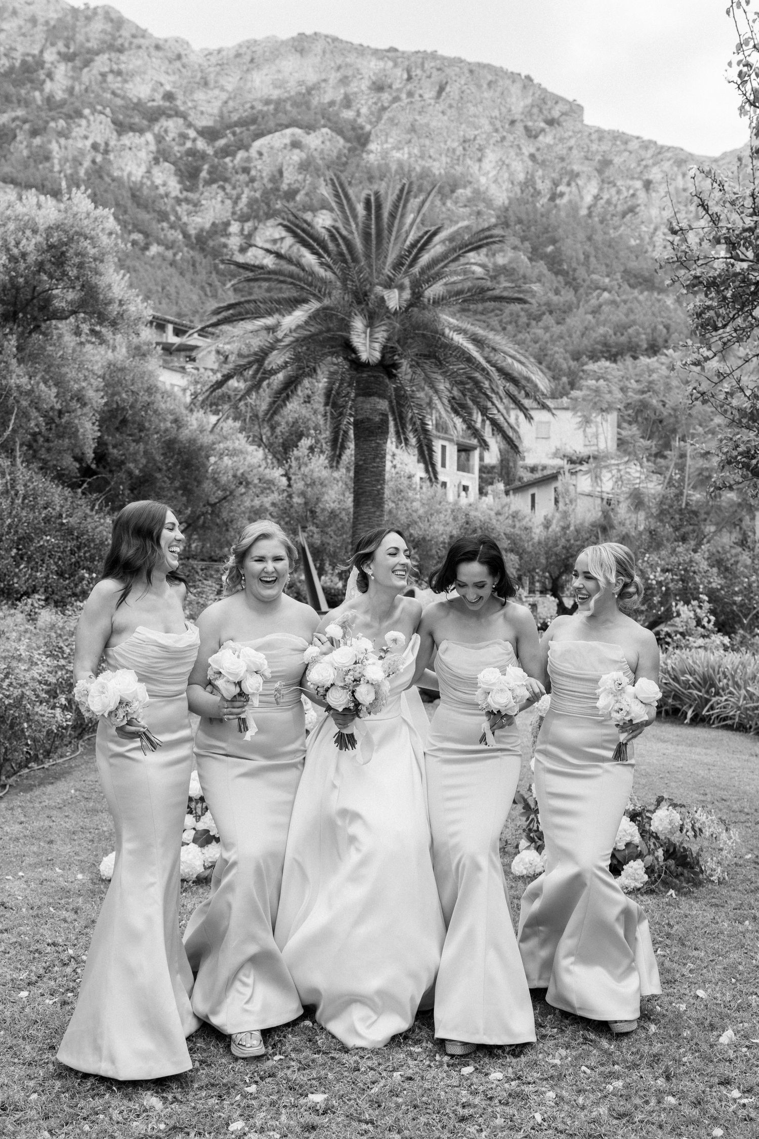 Bridesmaids in elegant white dresses pose together on garden lawn with mountain backdrop.