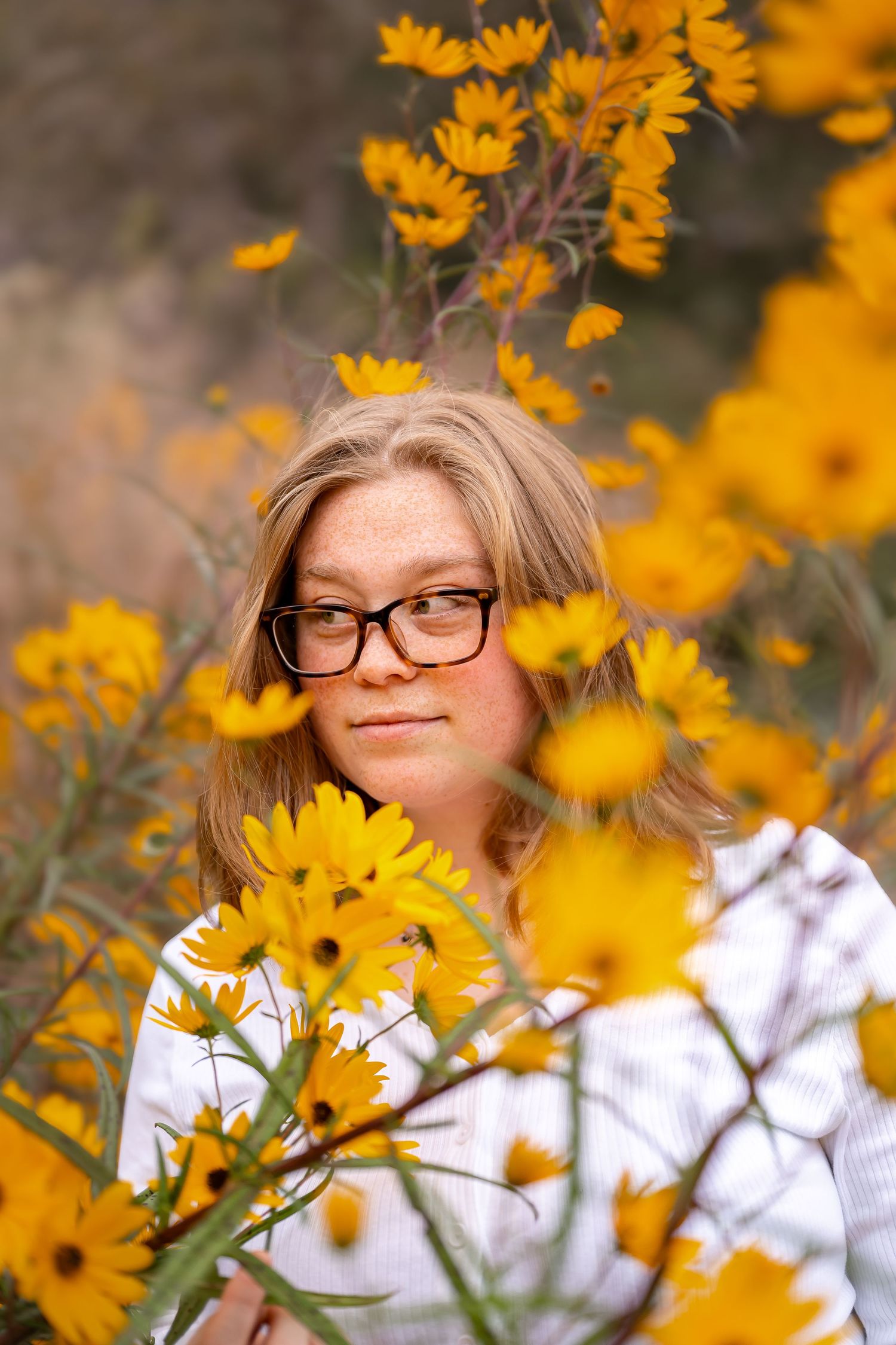Woman with glasses surrounded by yellow wildflowers, looking to the side with a thoughtful expression. Candid out photography in Hampton Rds