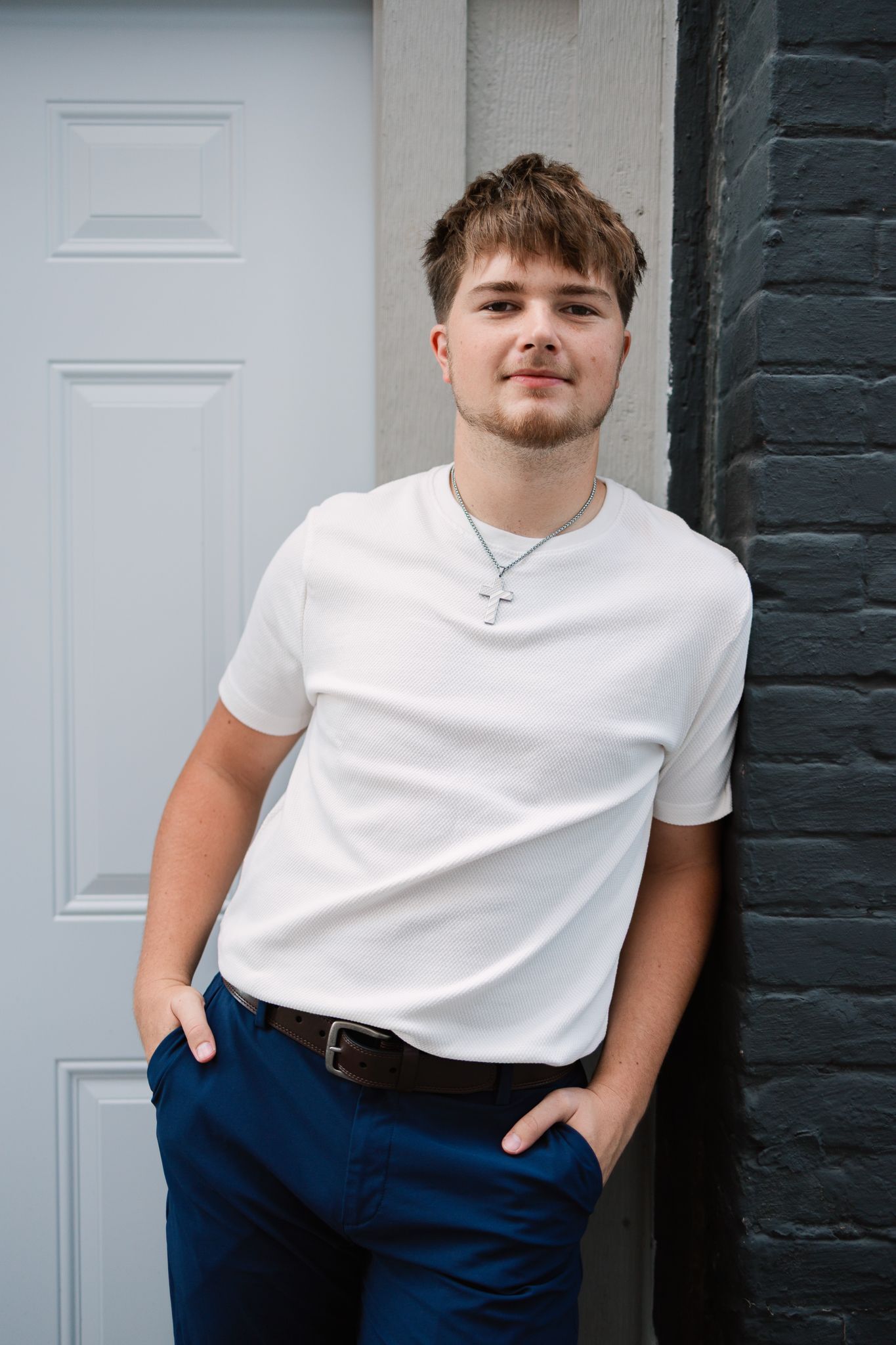 senior leaning against a dark gray brick wall and white door with his hands in his pockets