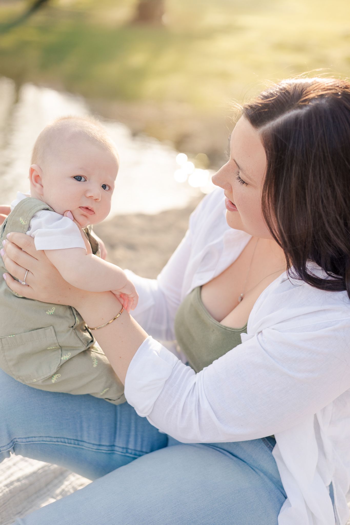 mother holding baby looking at him while sitting on a rock next to a creek
