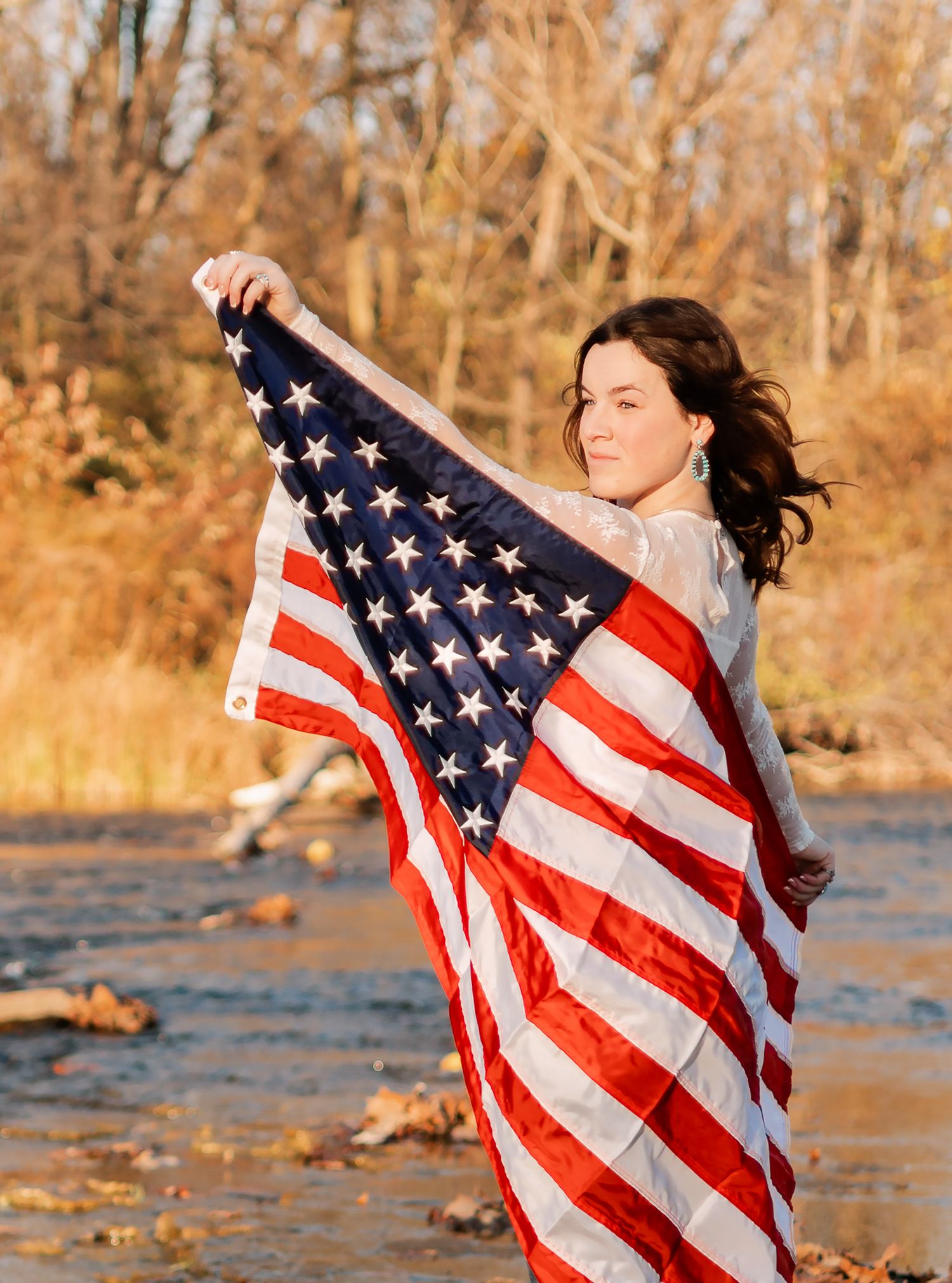 high school senior girl holding the American flag behind her, she is standing on rocks near water looking at the sunset in Pendleton, IN