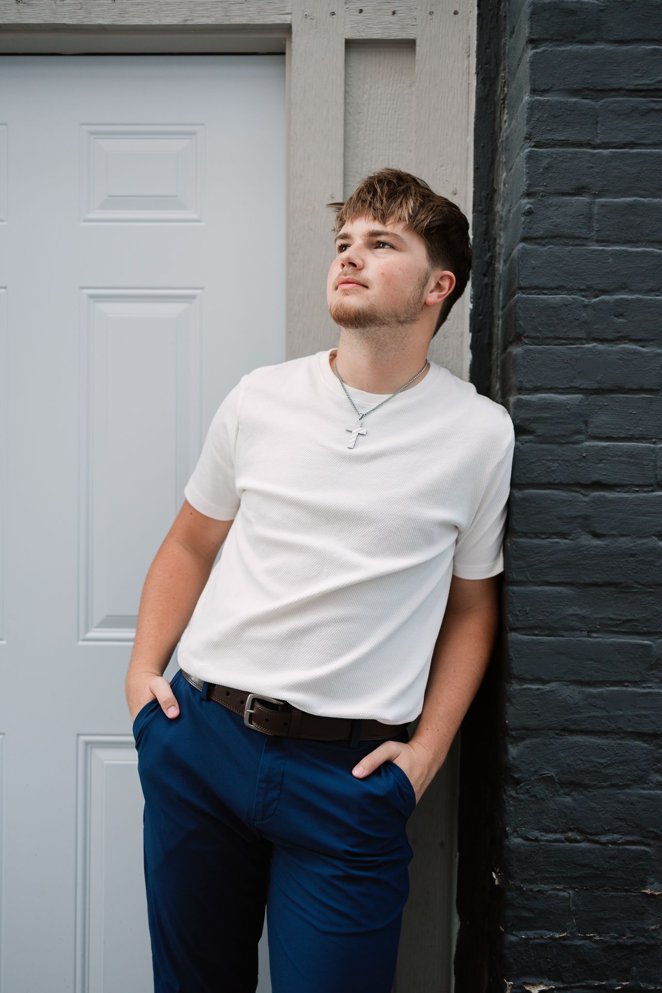 senior leaning against a dark gray brick wall and white door with his hands in his pockets, looking away