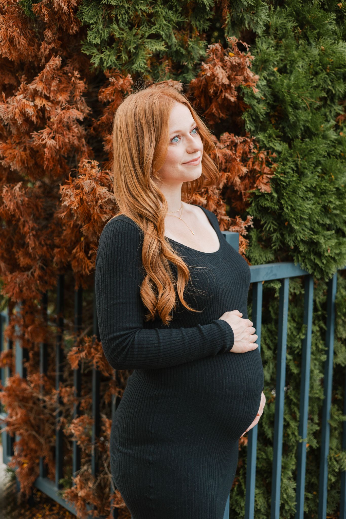 fall maternity mom to be standing in front of a brown and green bush wearing black and holding her baby bump