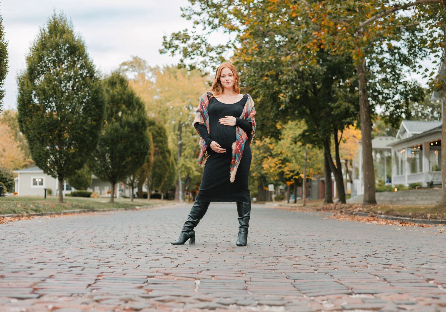 Fall maternity session with mom to be standing on a brick road wearing a black dress and boots with a plaid scarf