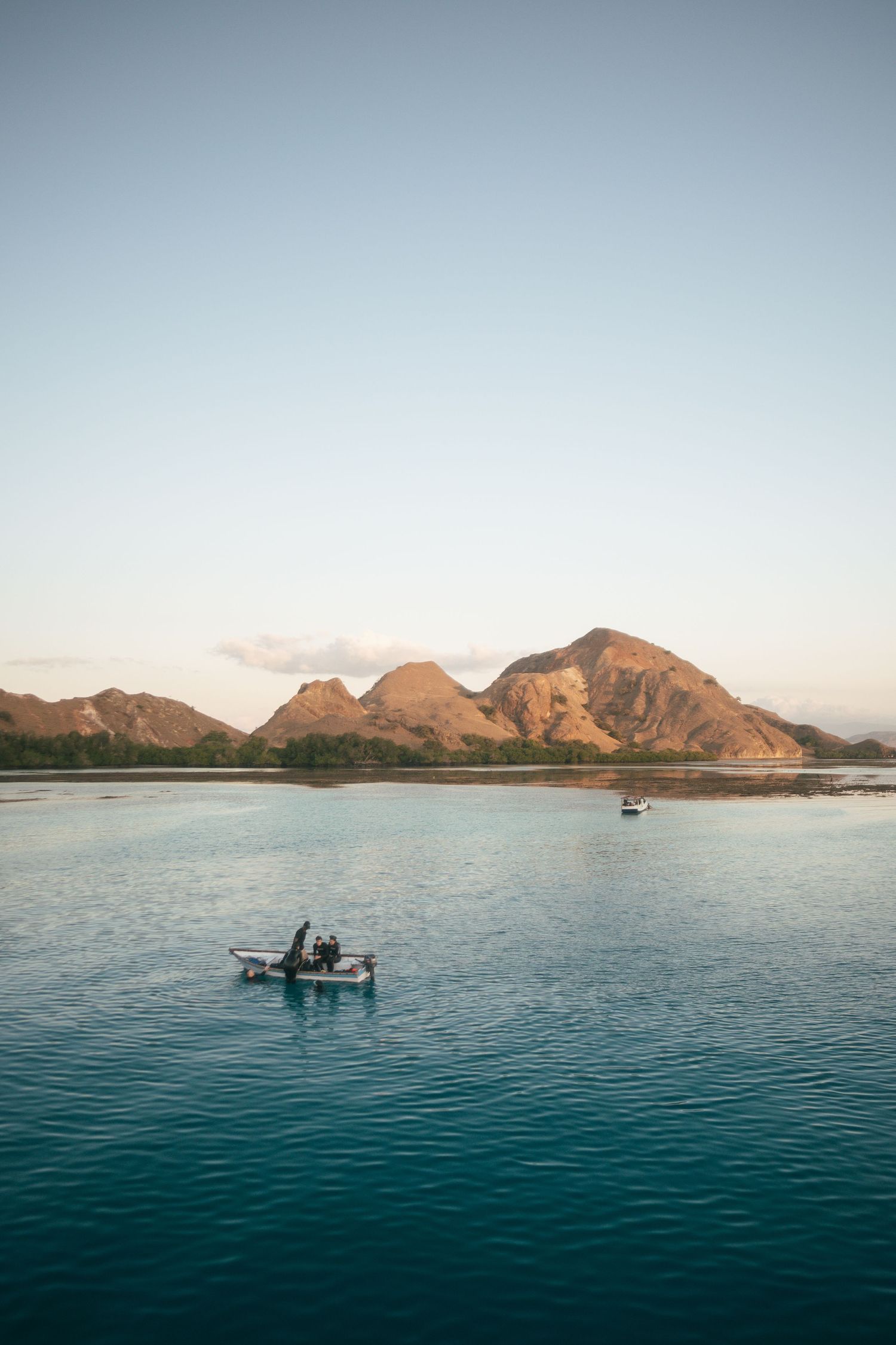 Small boats float on calm turquoise waters with rugged mountains rising along the coastline at sunset.