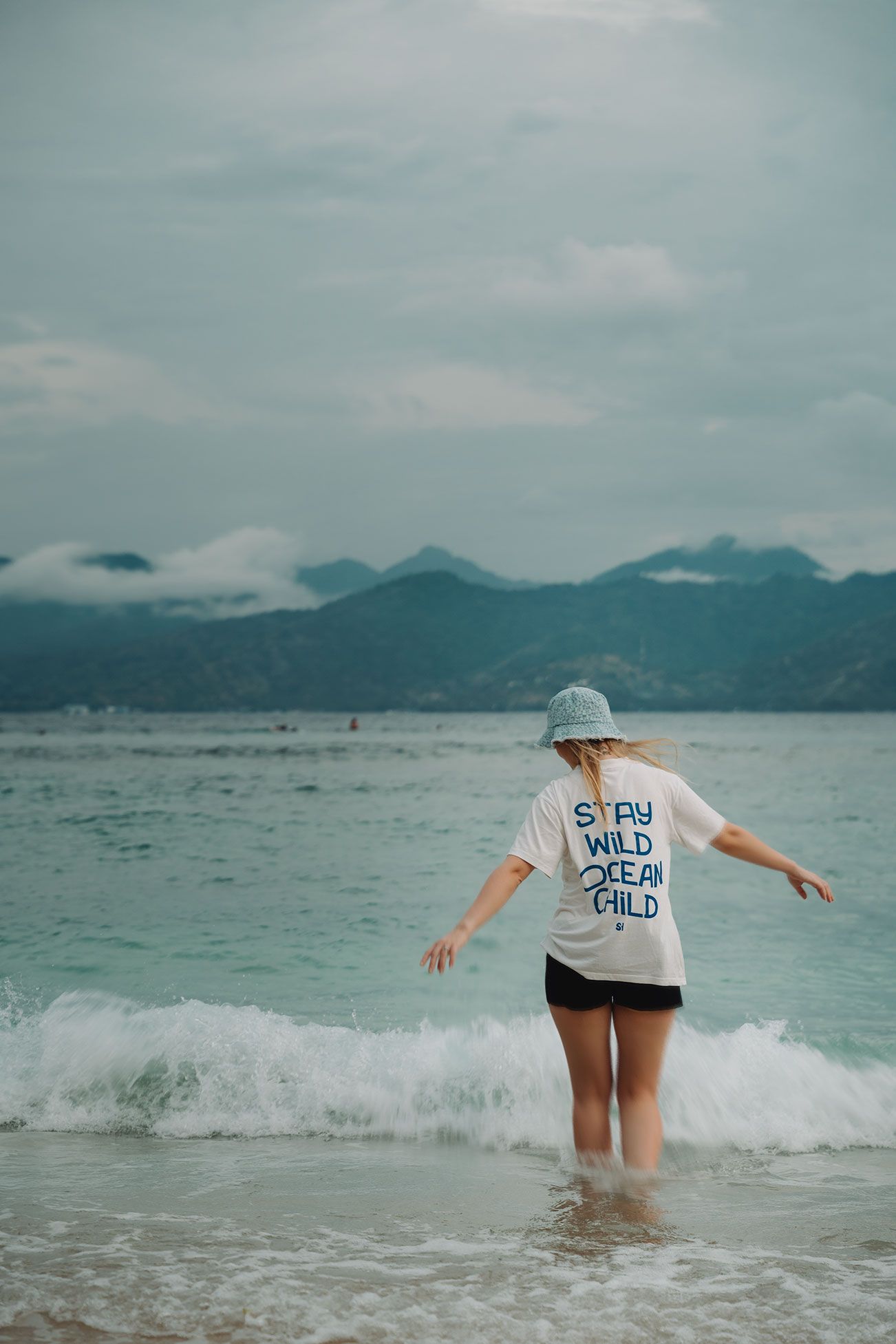 Person wearing 'Ocean Child' shirt playing in ocean waves with mountains in background.