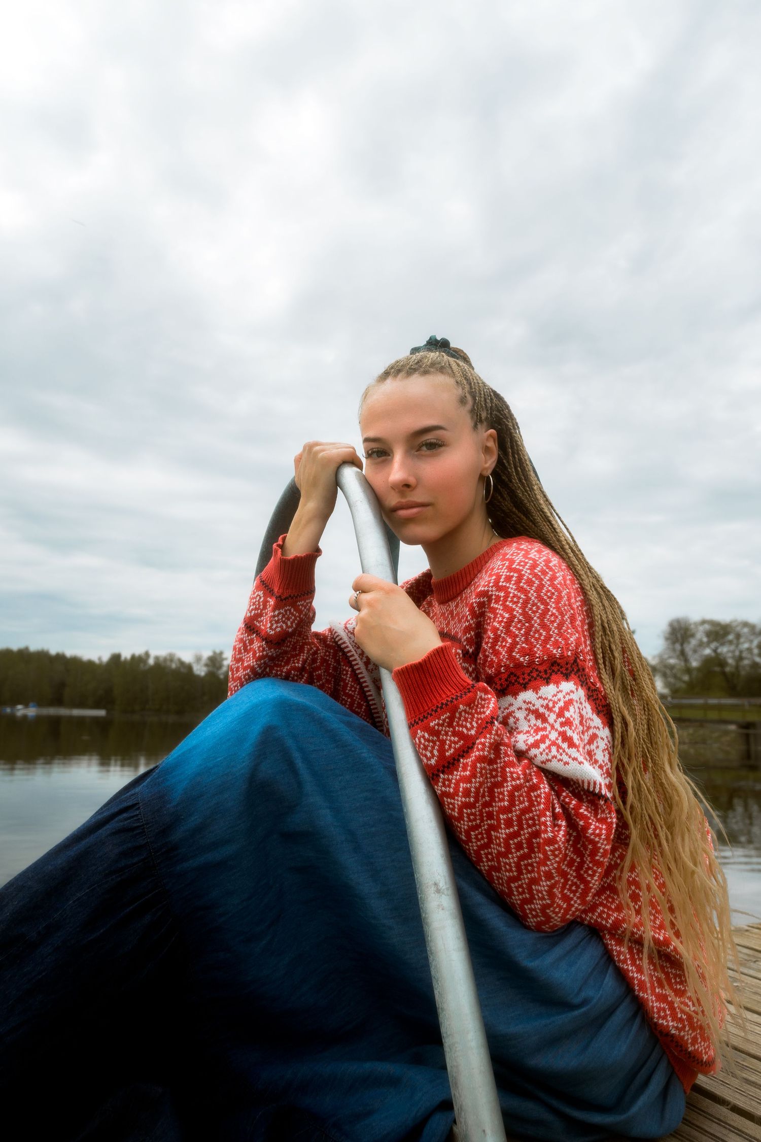 Person in red knit sweater reclining on dock by lake under cloudy sky.