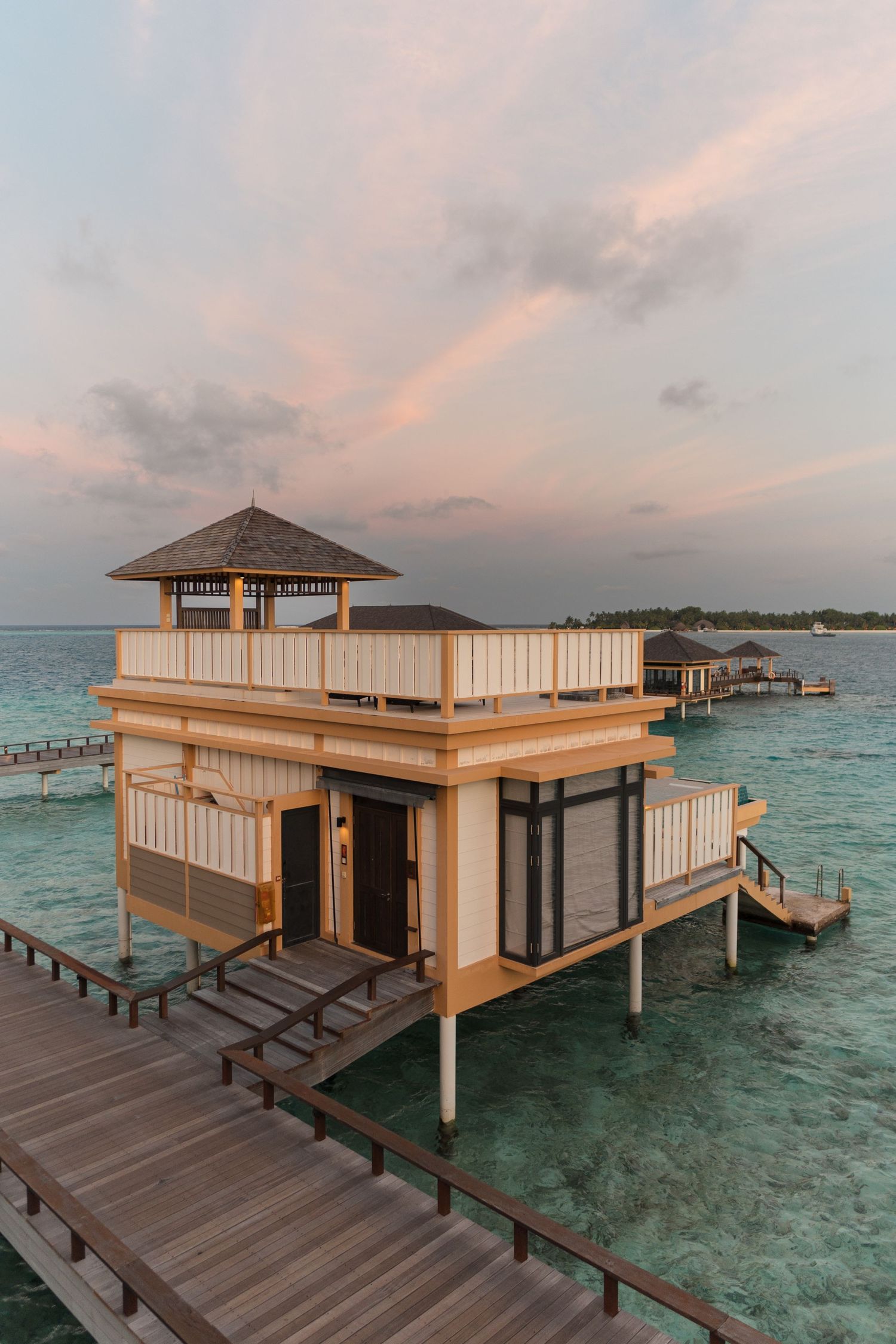 Luxury overwater bungalows with wooden decks reflecting pink sunset colors over tropical waters.