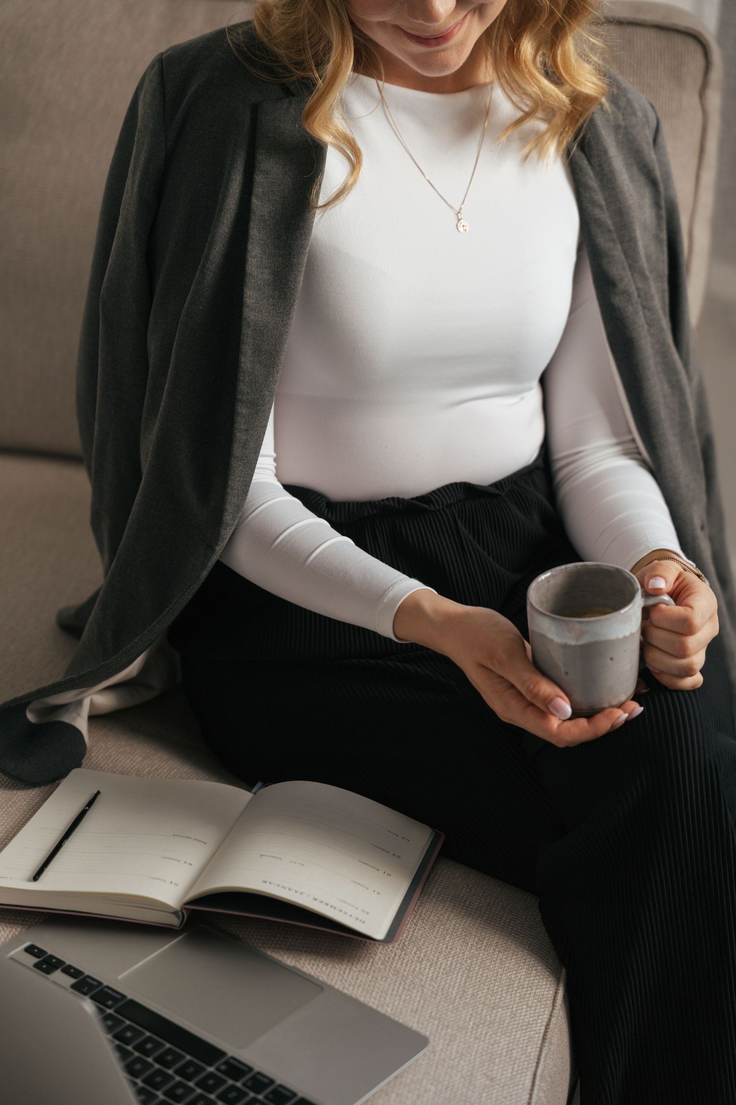 Person in gray cardigan and white top working with laptop and coffee.