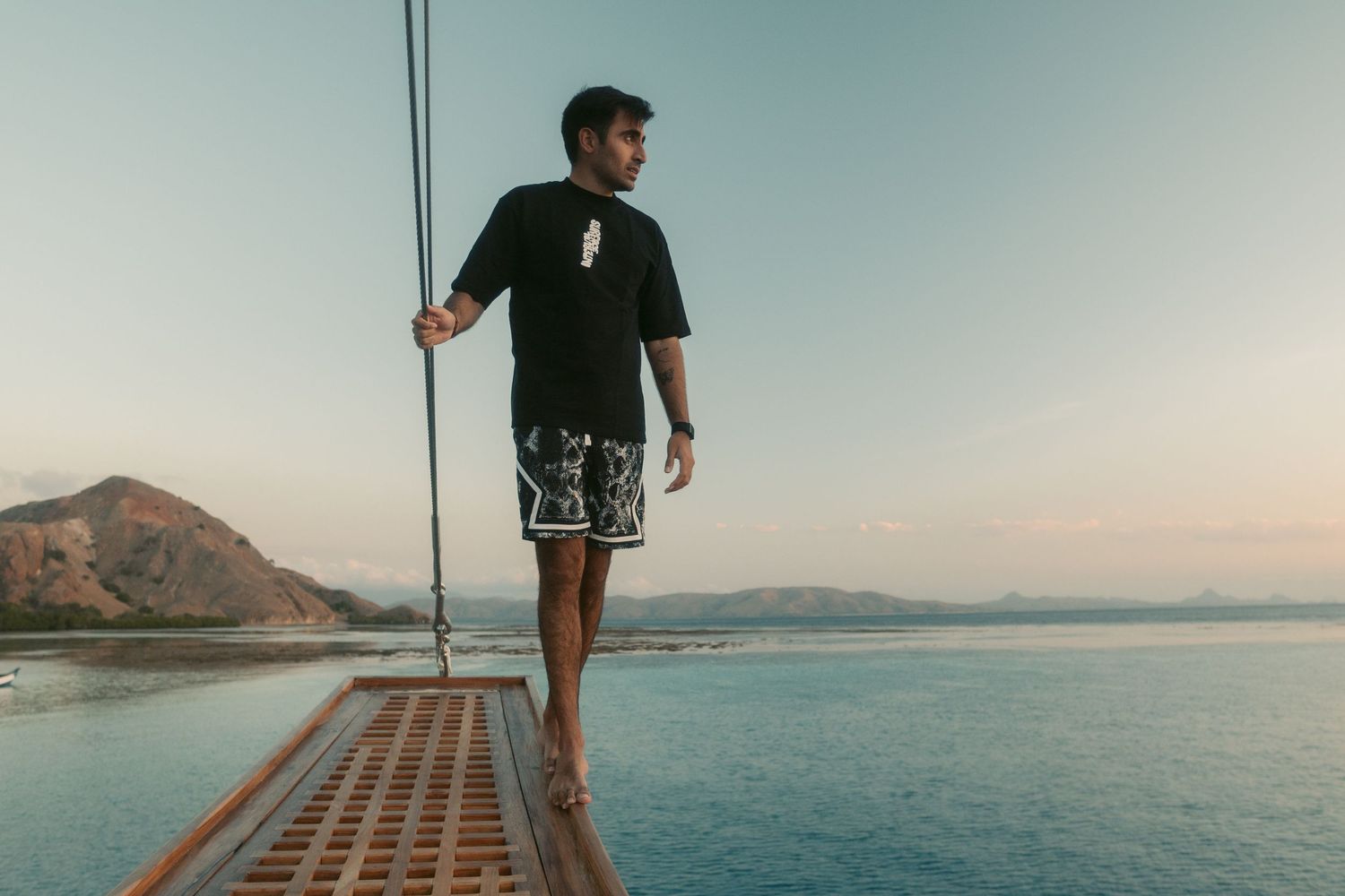 Person walking on a suspended bridge with ocean and mountain views at sunset.